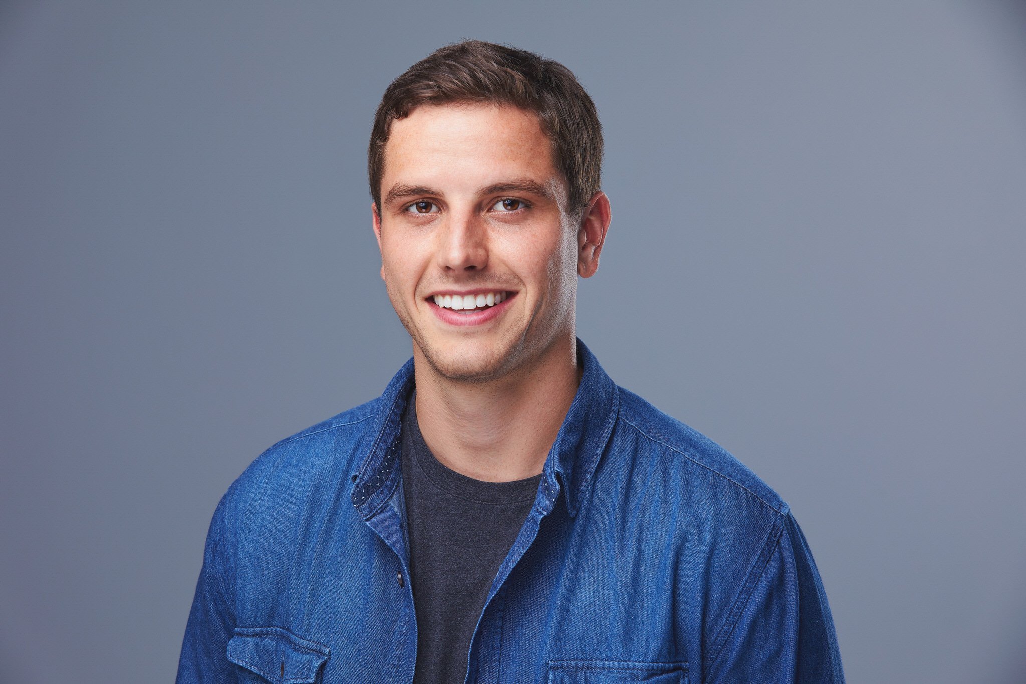 Professional headshot of Ray in a blue shirt against a soft grey studio background.