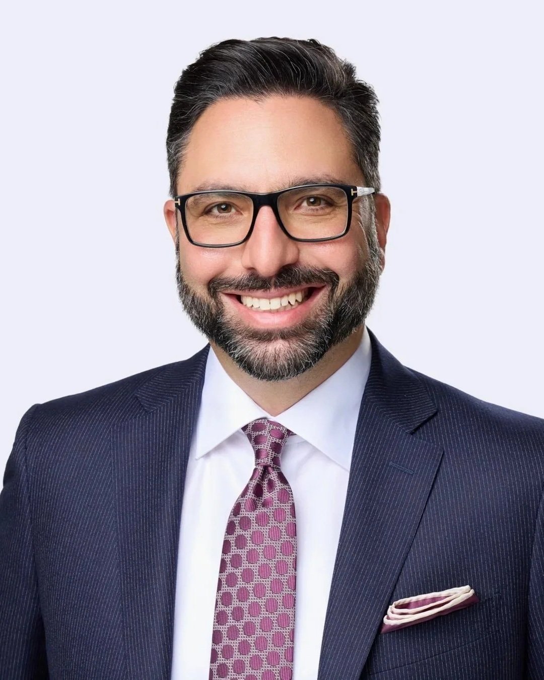 Professional business headshot of a smiling man in glasses and a navy suit against a light grey studio background.