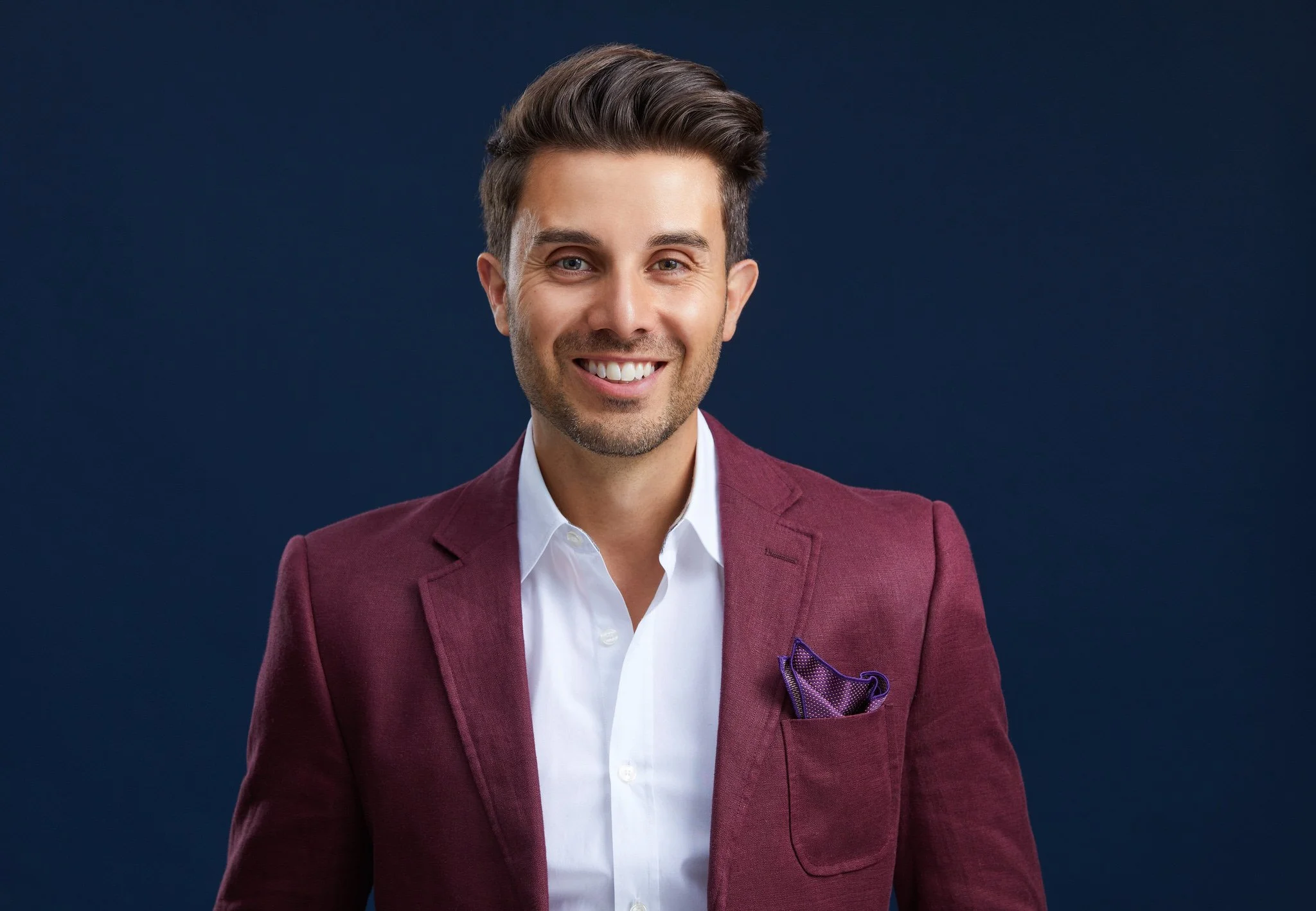 Contemporary business headshot of a smiling man in a burgundy jacket against a navy studio backdrop.