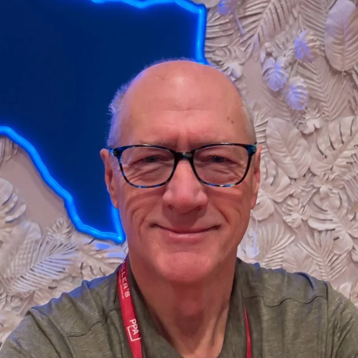 Bald man with glasses and a red lanyard in front of a textured wall with neon lighting.
