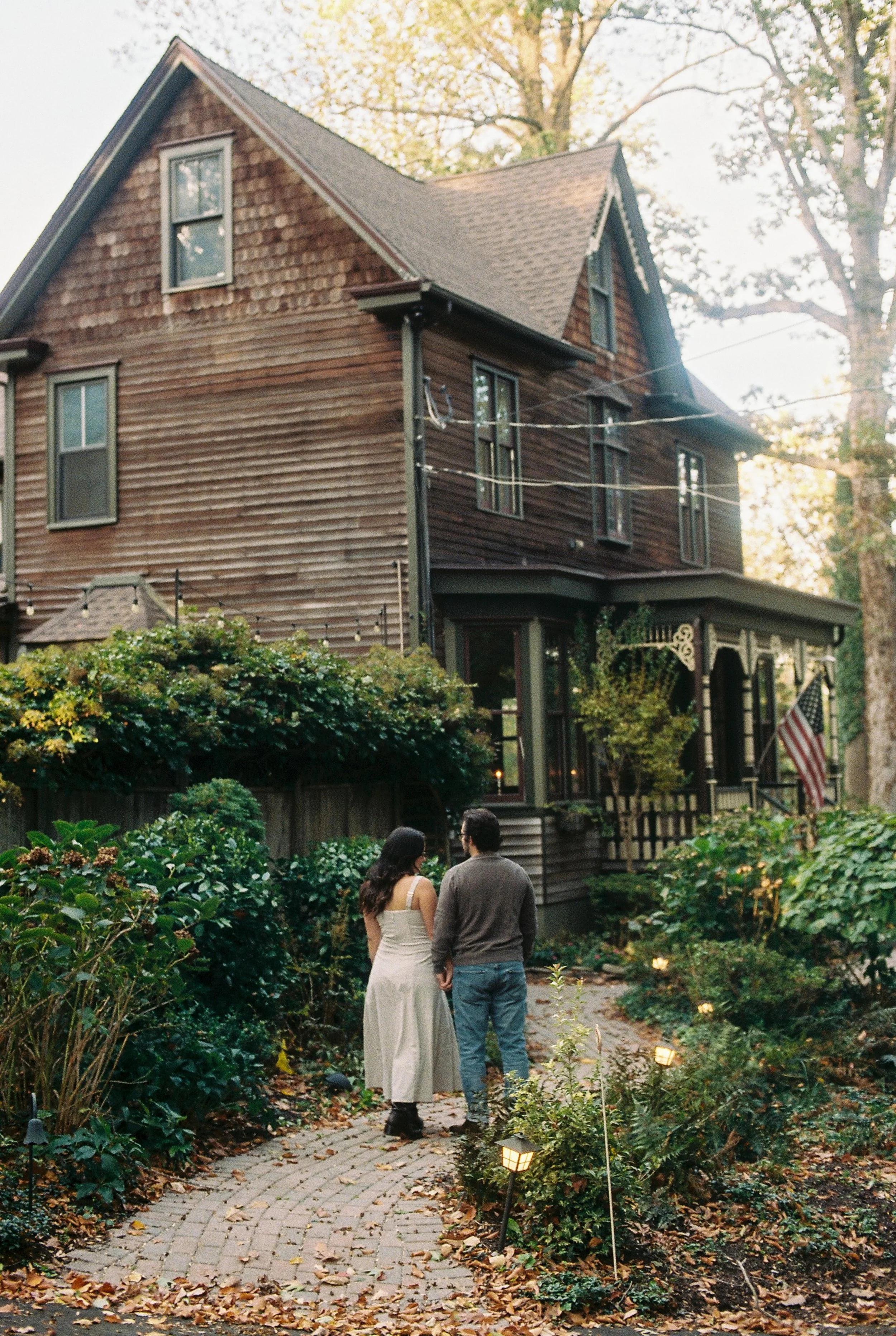A couple walking hand in hand on a brick pathway in a garden, with a large wooden house in the background, surrounded by trees and turn-on garden lights.