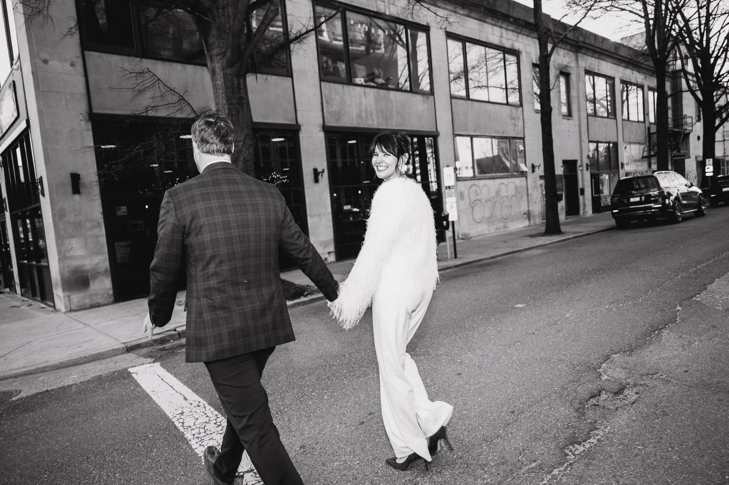 A woman in white pants and a fluffy top holding hands and smiling at a man in a checkered jacket walking across the street in an urban setting with shops and cars.