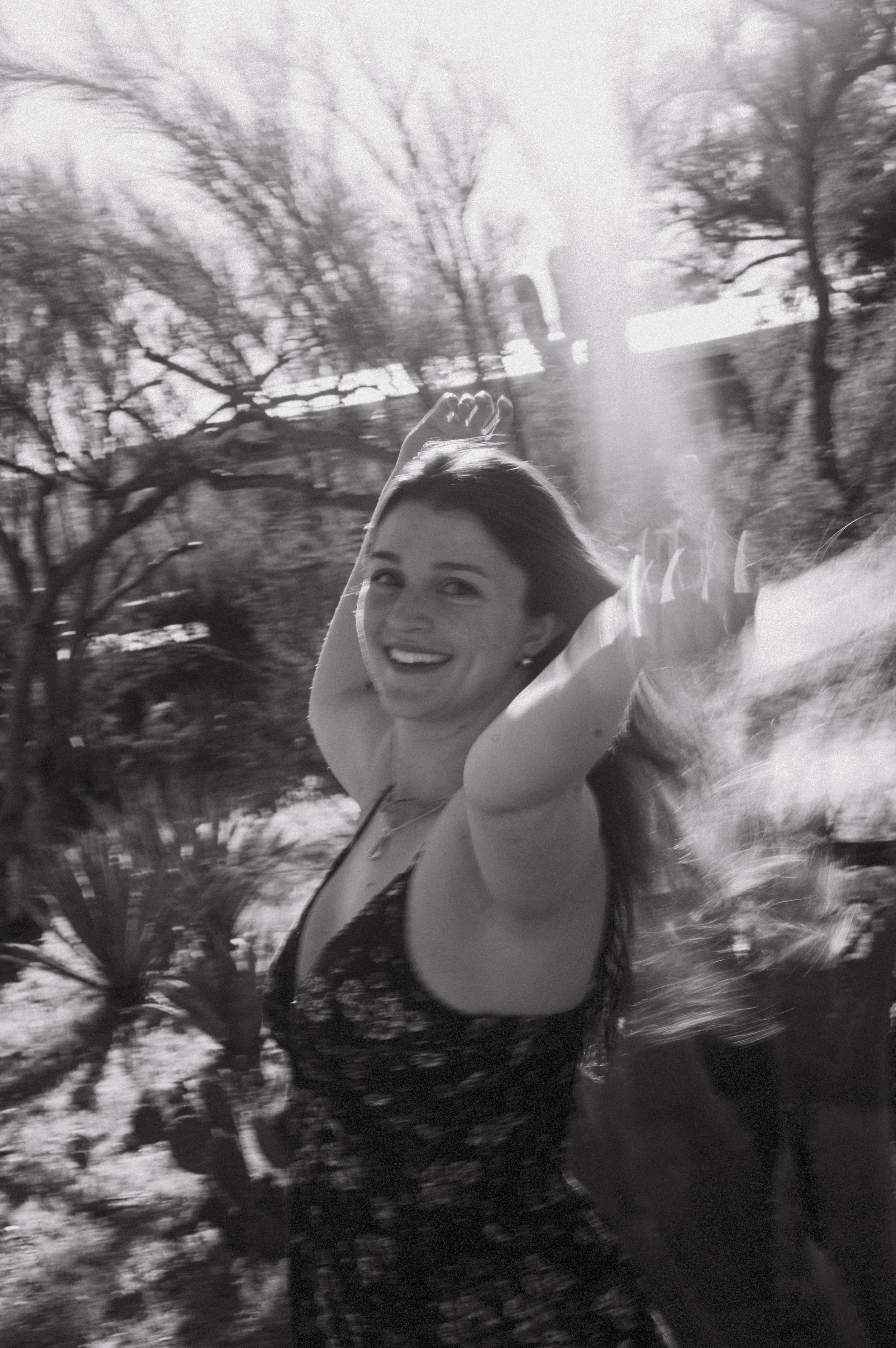 A woman smiling and raising her arms outdoors with blurred trees and sunlight in the background.