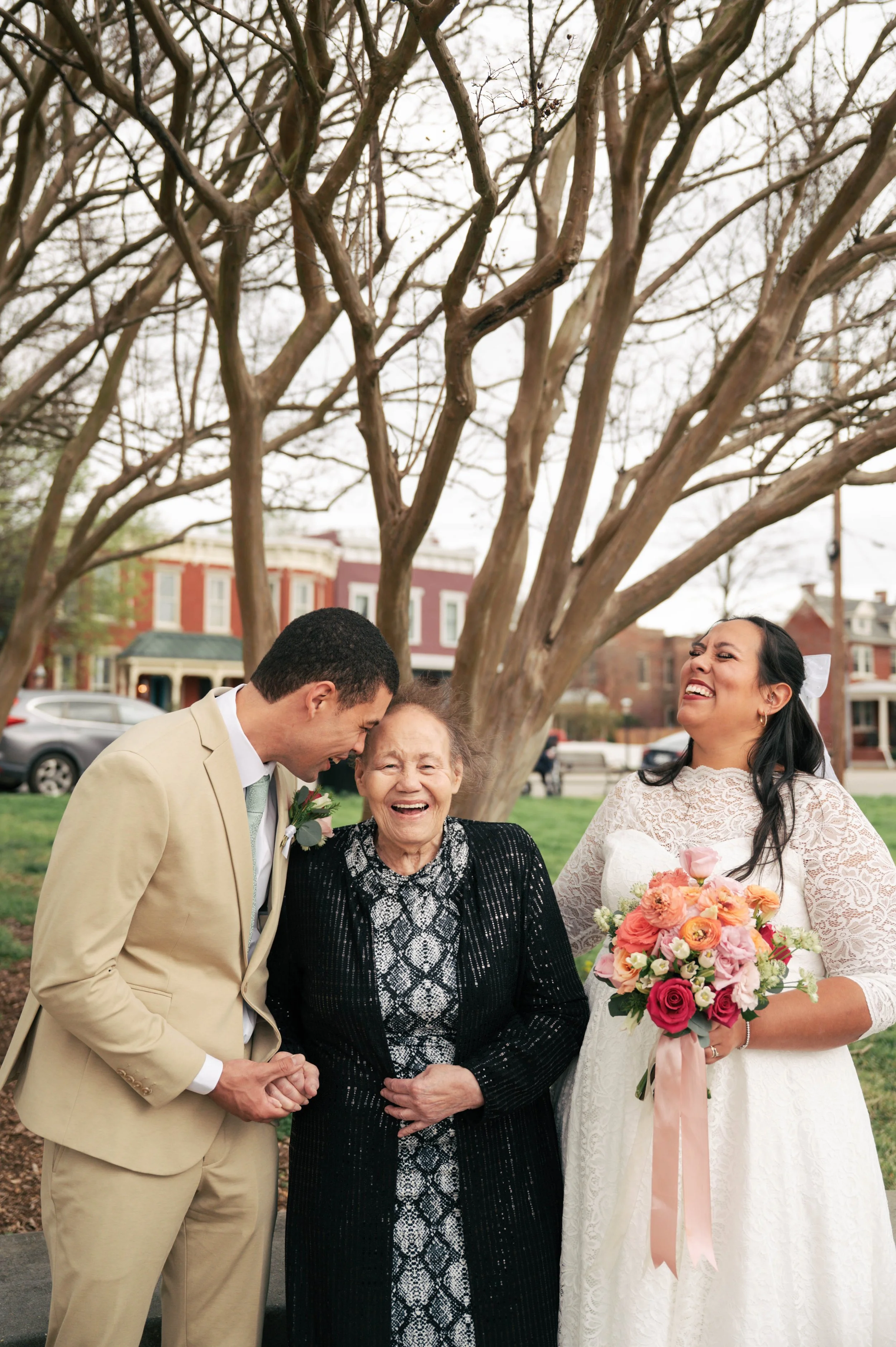 A wedding celebration featuring three women standing outside under a tree, sharing a joyful moment. The woman on the right is holding a bouquet of flowers and is wearing a white lace wedding dress, smiling and laughing. The woman in the middle, an elderly woman, is wearing a black and gray patterned dress with a black cardigan, also smiling. The man on the left, dressed in a beige suit, is leaning toward and sharing a laugh with the elderly woman.