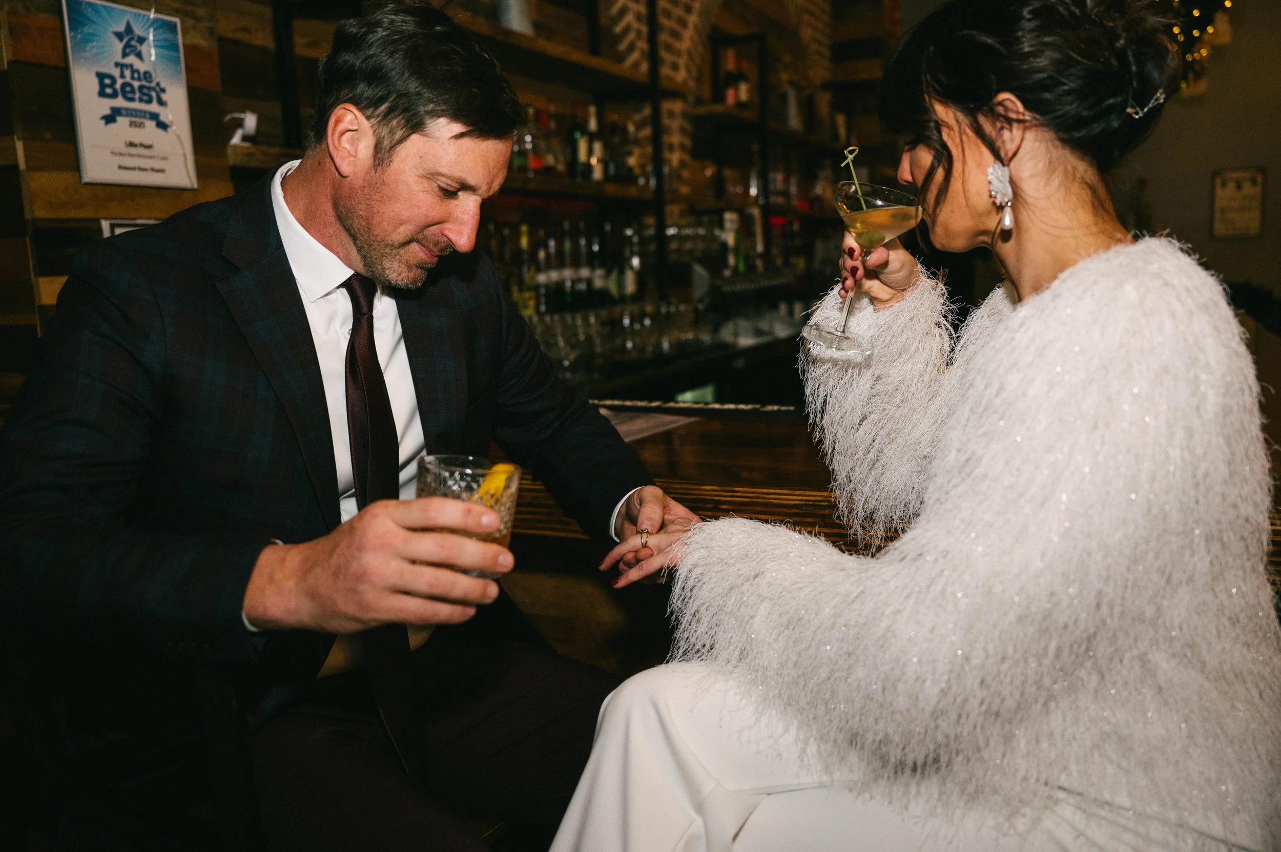 A man in a suit holding a cocktail and a woman in a white sparkly dress with jewelry sitting at a bar, holding hands, and enjoying drinks in a cozy bar setting.