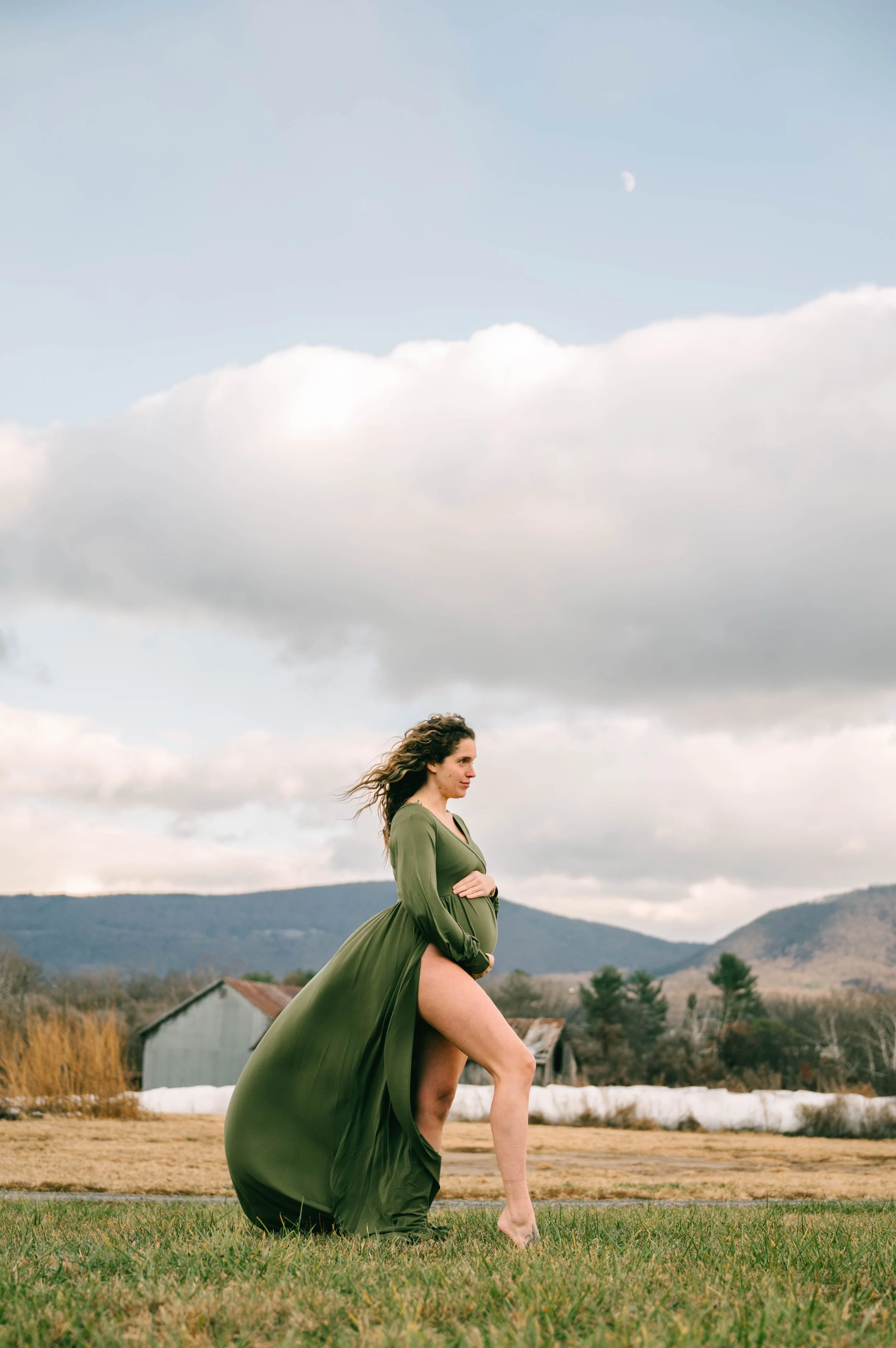 A pregnant woman in a flowing green dress stands outdoors on a grassy field with a rural landscape and hills in the background, under a partly cloudy sky.