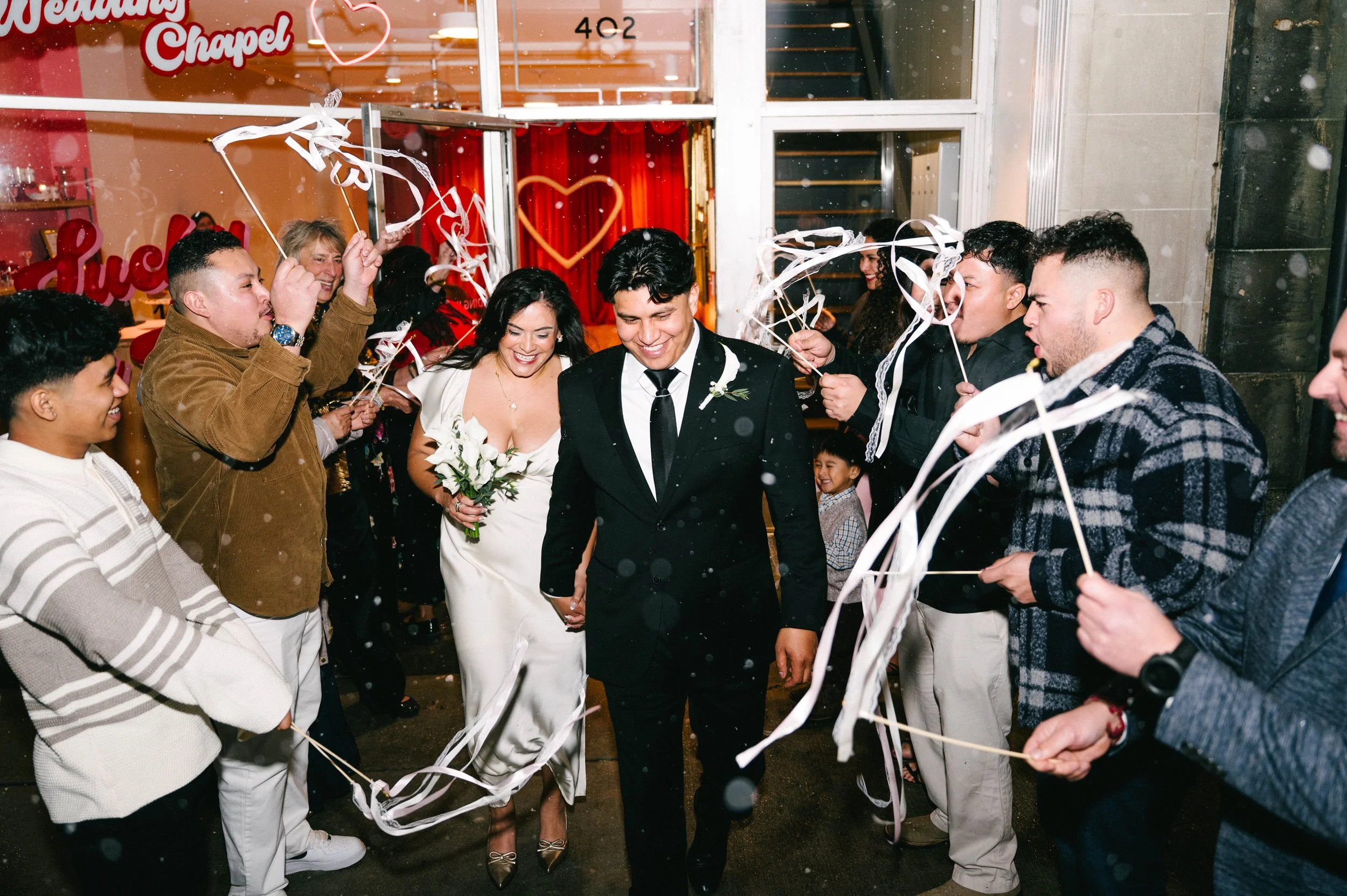 A bride and groom walking through a crowd of guests holding ribbons and streamers outside a wedding chapel, celebrating.