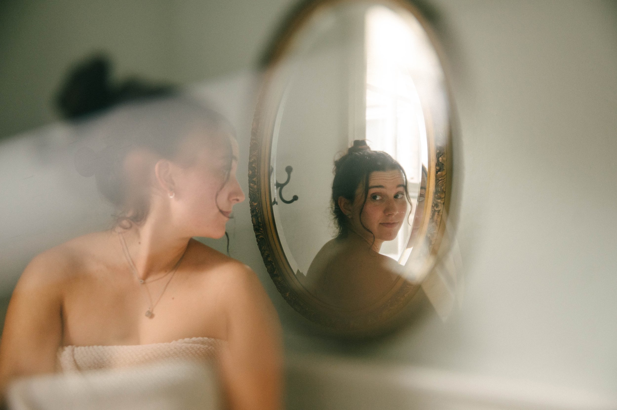 A woman looking at her reflection in an oval mirror, with her hair tied up, wearing a strapless top, in a softly lit room.