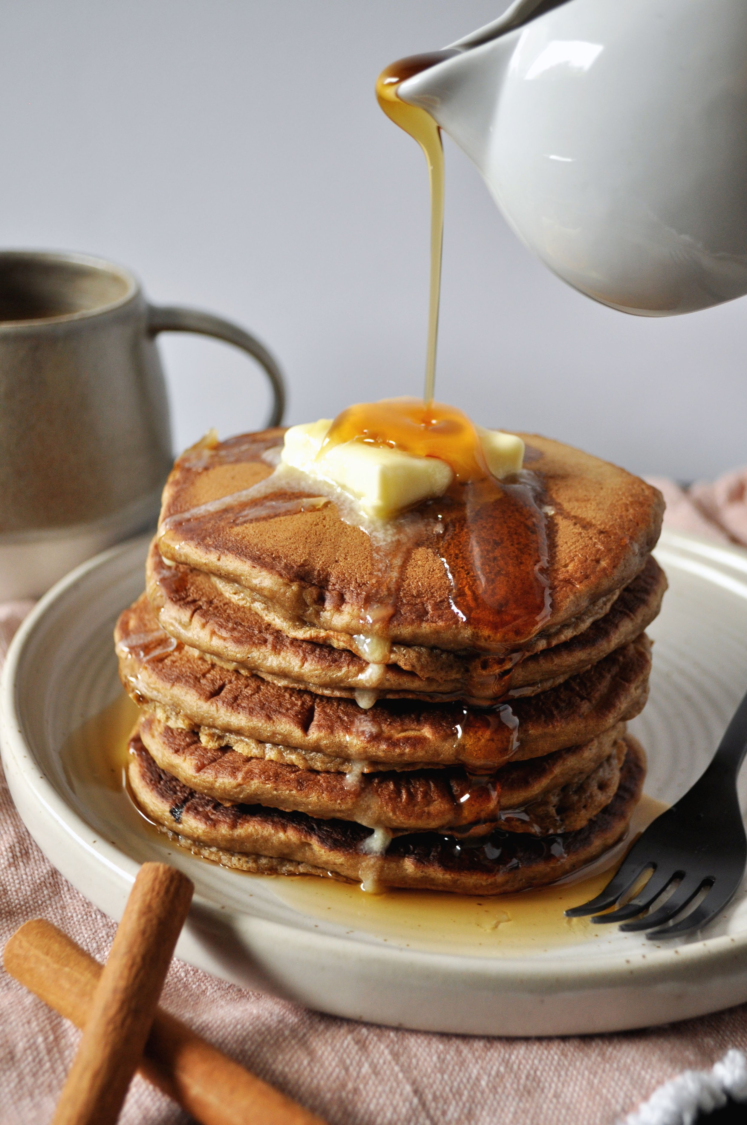 Stack of pancakes with butter and maple syrup being poured from a white pitcher, served in a ceramic plate with a dark fork and cinnamon sticks nearby. A mug is in the background.