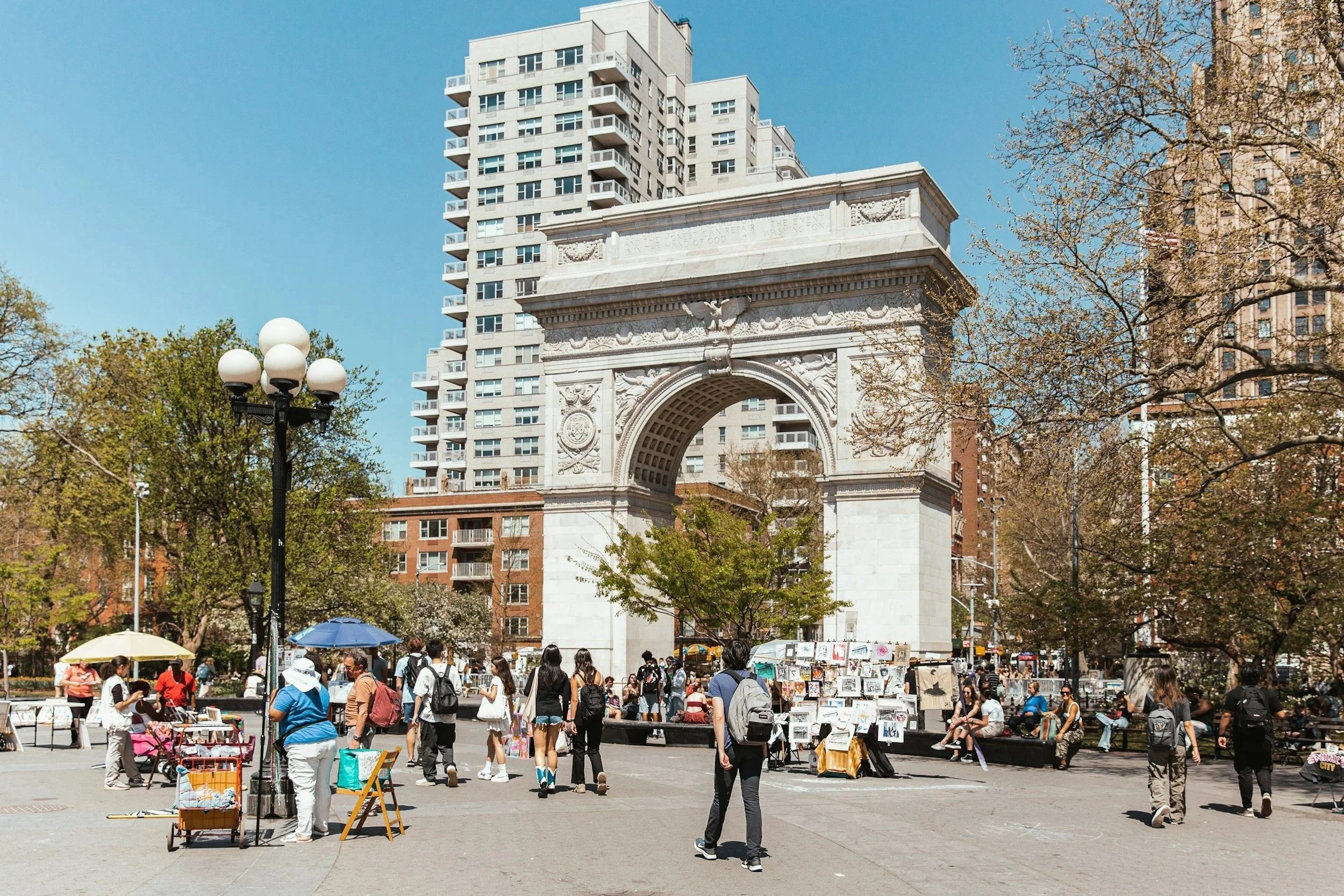 People walking and shopping at an outdoor market in front of the Washington Square Arch in New York City, with high-rise buildings and trees around.