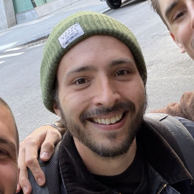 Close-up of a smiling man wearing a green beanie and black jacket, with part of two other people visible on either side, outdoors.