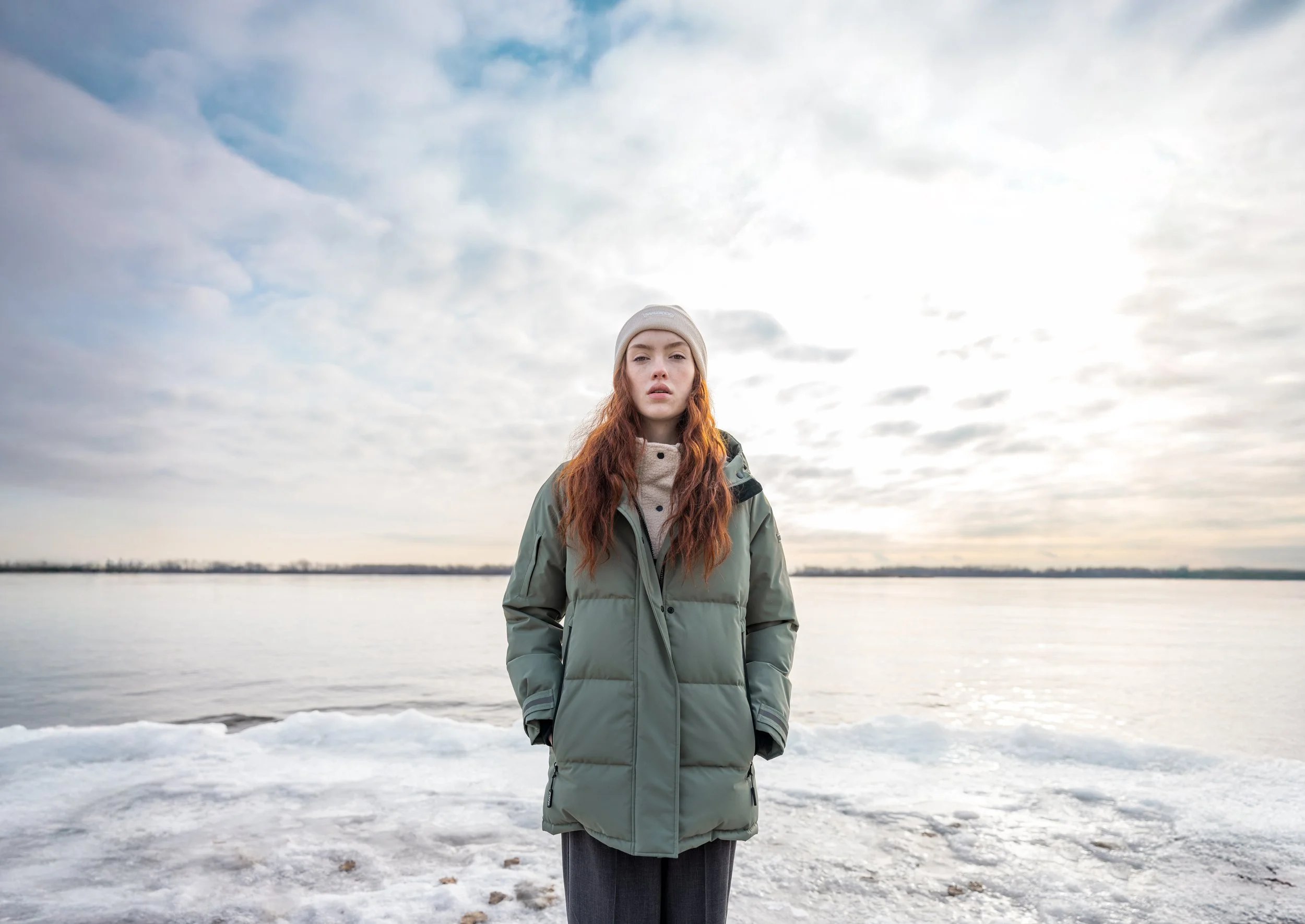 A young woman with long red hair wearing a beige beanie and green winter coat standing on snowy ground near a body of water during cloudy weather.