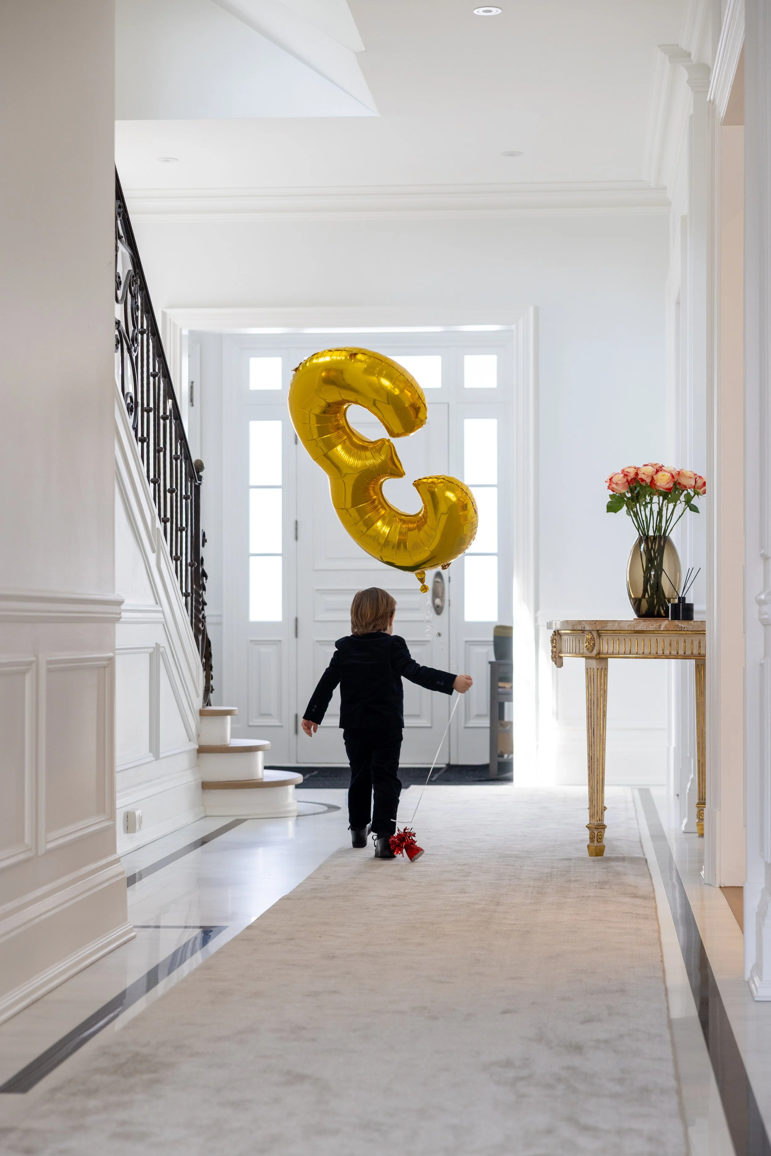 A young boy dressed in a black suit walking in a bright, elegant foyer, holding a balloon shaped like the number three. The foyer features a cream-colored rug, white walls, a gold-colored table with pink roses, and a large golden number '3' balloon f