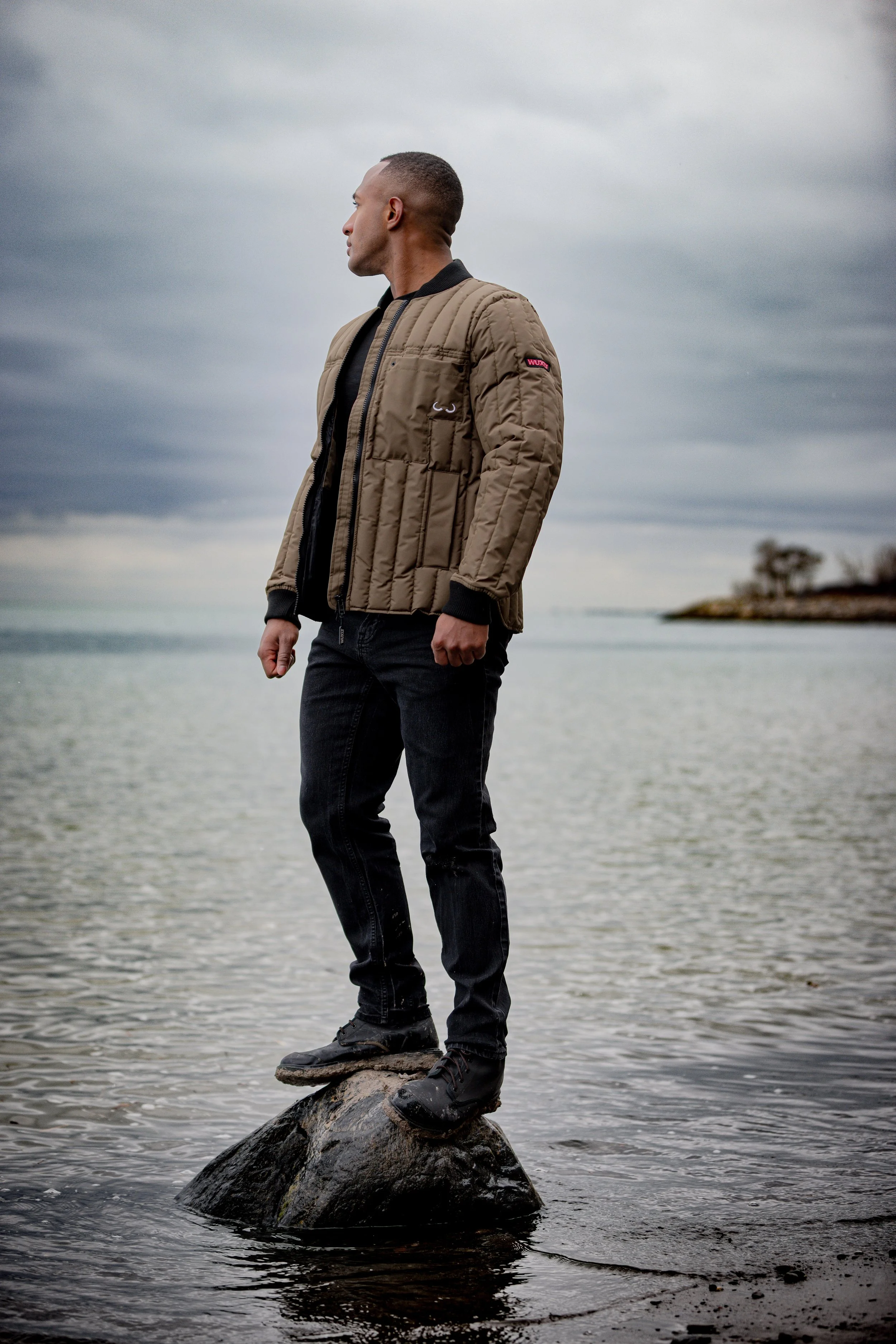 A man stands on a rock in a body of water with an overcast sky in the background, facing left, wearing a tan quilted jacket, black jeans, and black shoes.