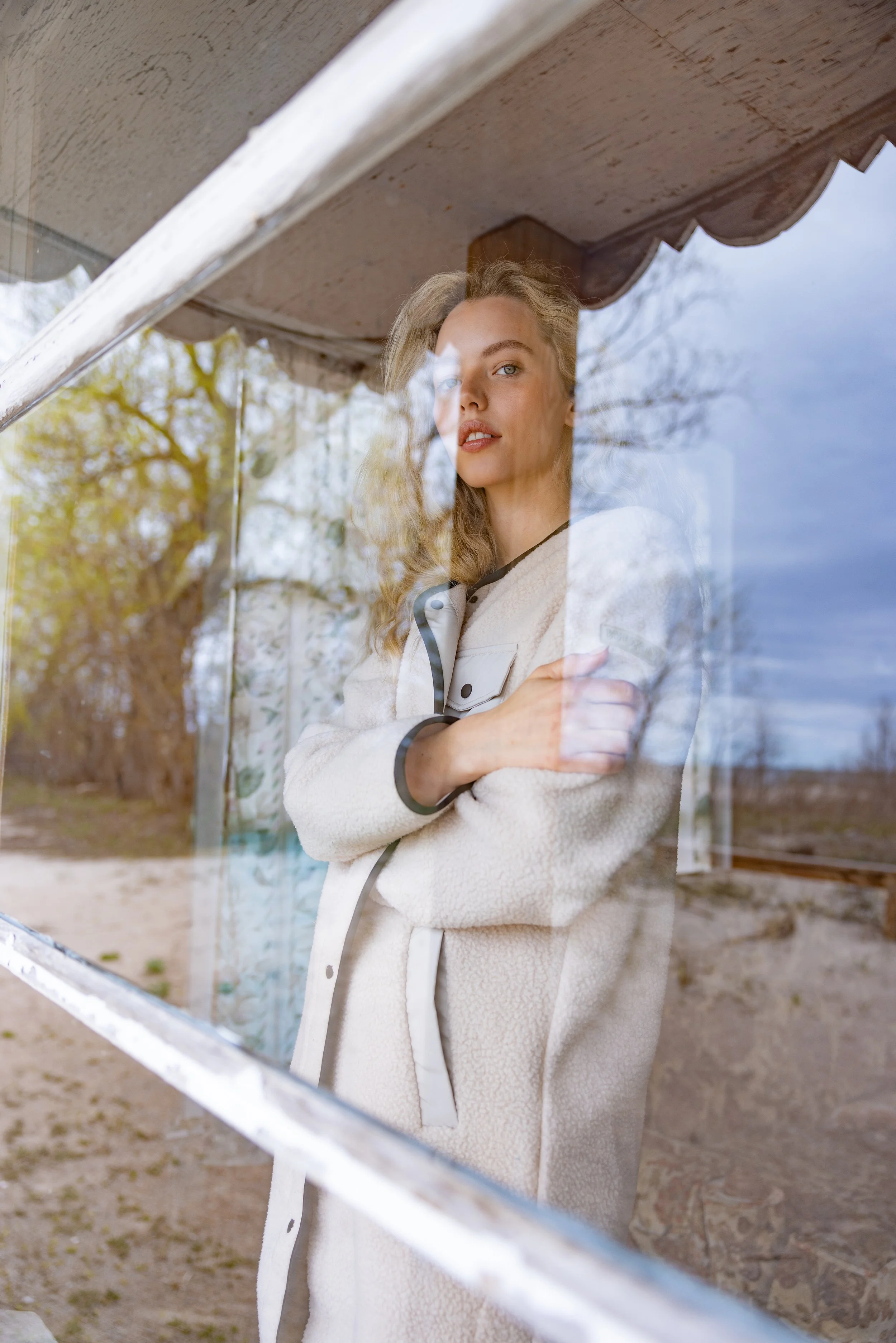 A young woman with wavy blonde hair wearing a beige coat and standing behind a glass window, reflecting trees and a cloudy sky outside.
