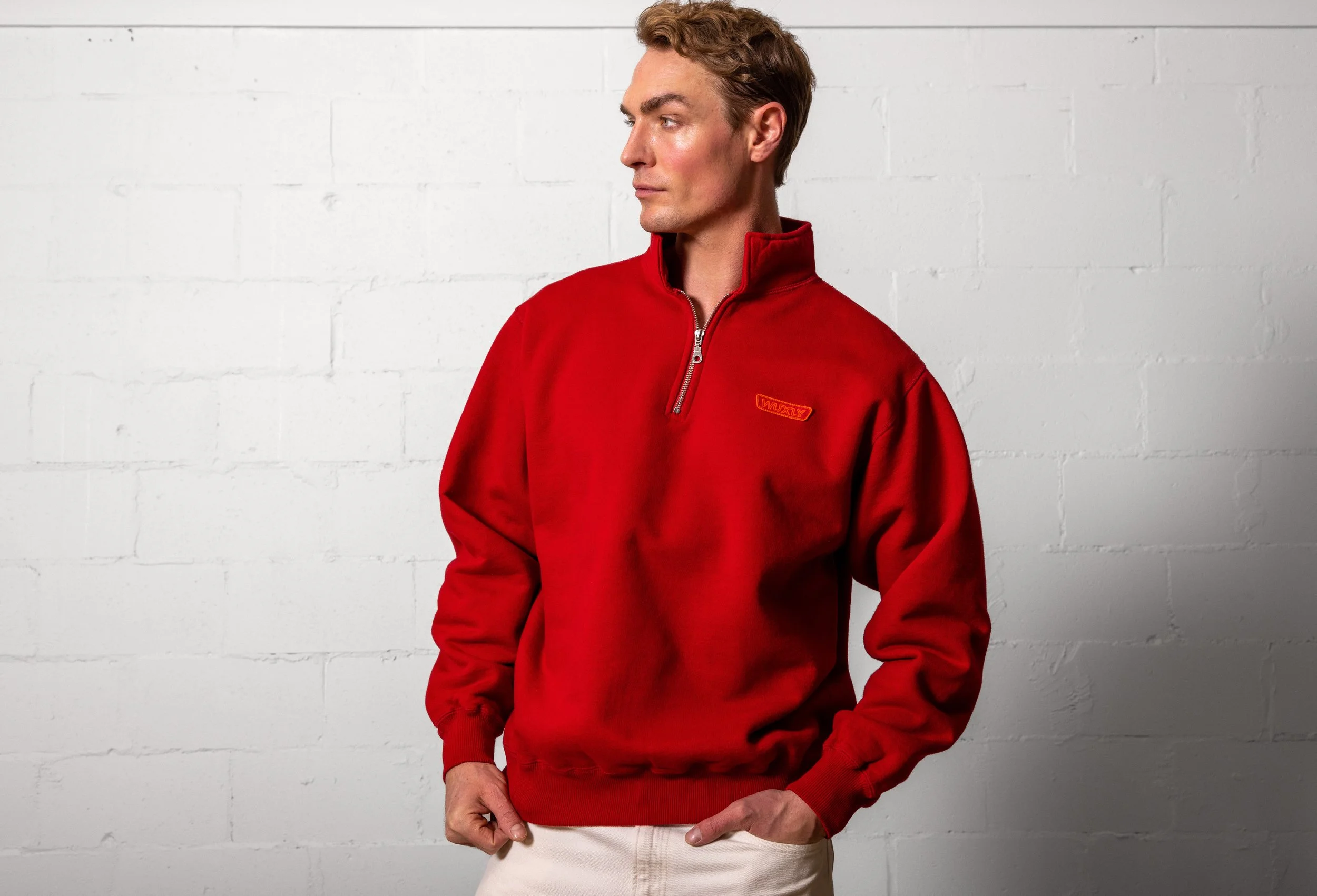 Man wearing a red quarter-zip fleece pullover with a logo on the chest, standing against a white brick wall