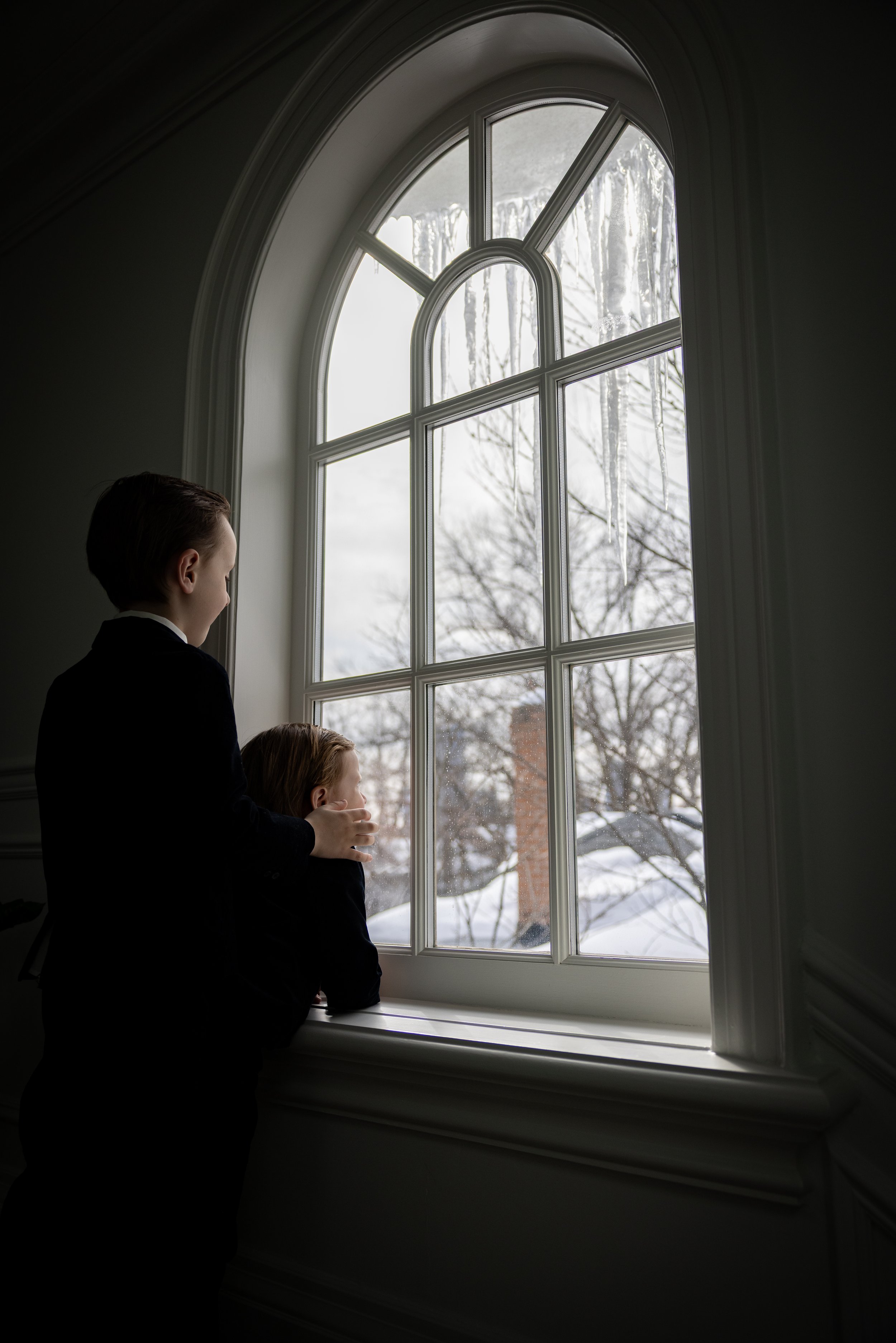 Two children looking out a large arched window with icicles hanging outside, winter scene.