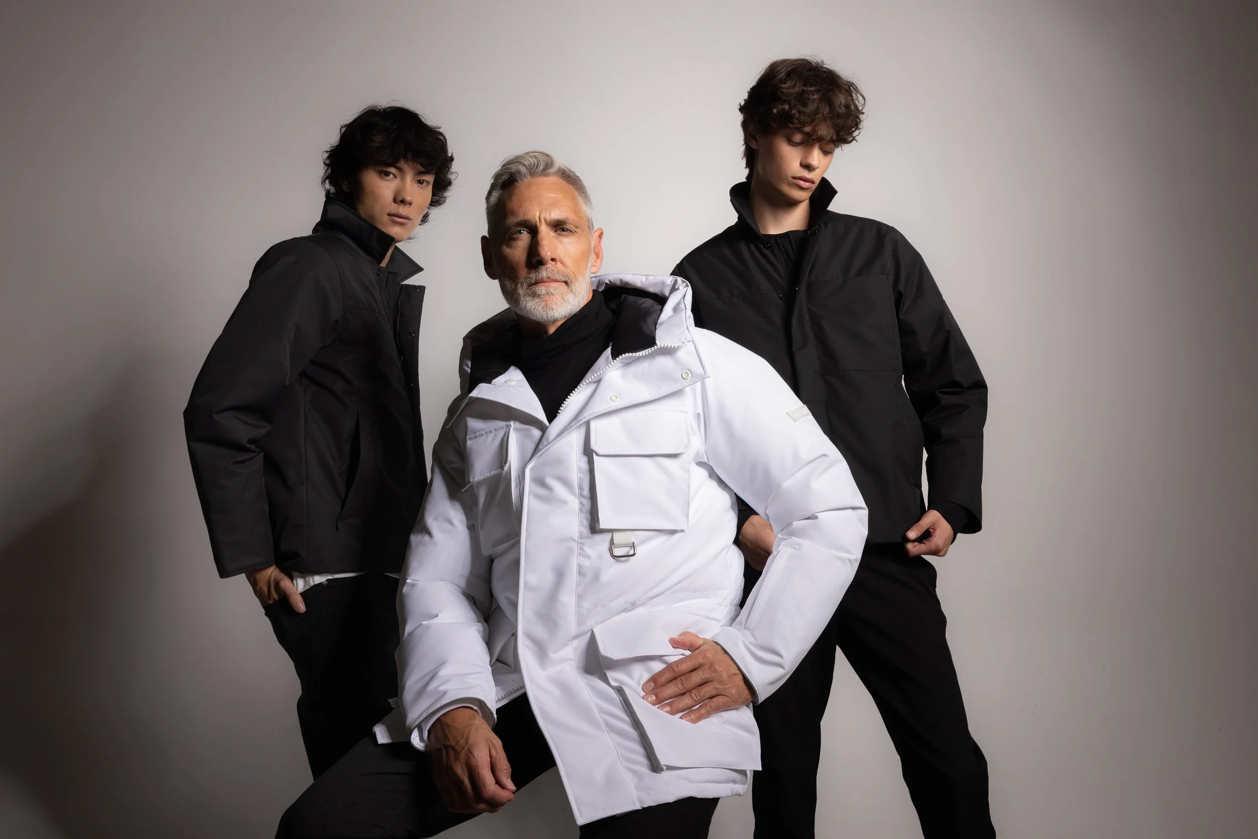 Three models pose against a plain background, with the elderly man in a white jacket in the center and two young men in black jackets flanking him.
