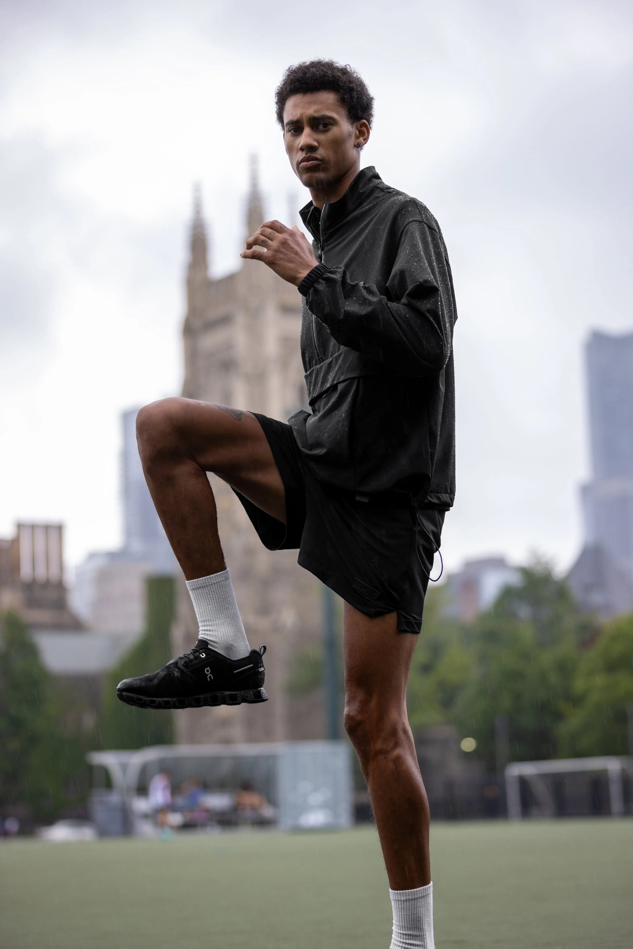 A young man wearing a black rain jacket, black shorts, white socks, and black running shoes stretches his leg on a sports field during rainy weather, with city buildings and a historic church tower in the background.