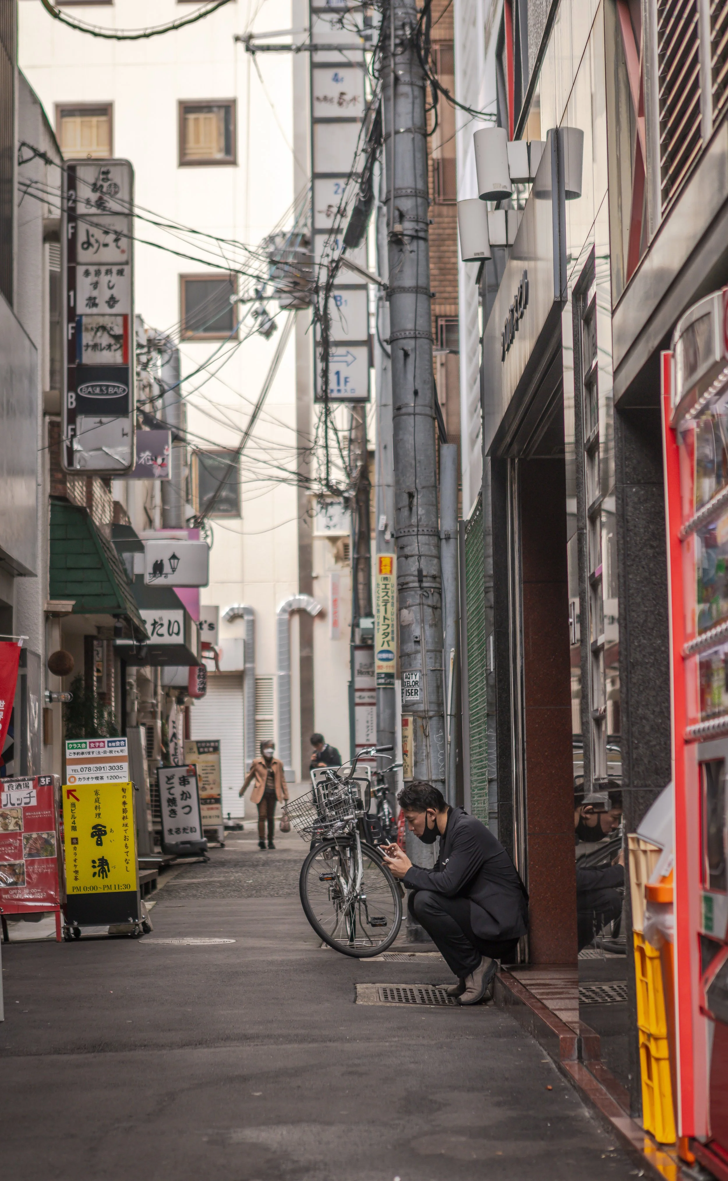 A man in a black suit sitting on a small step outside a building, looking at his phone, with a bicycle parked nearby on a narrow street with various signs and pedestrians.