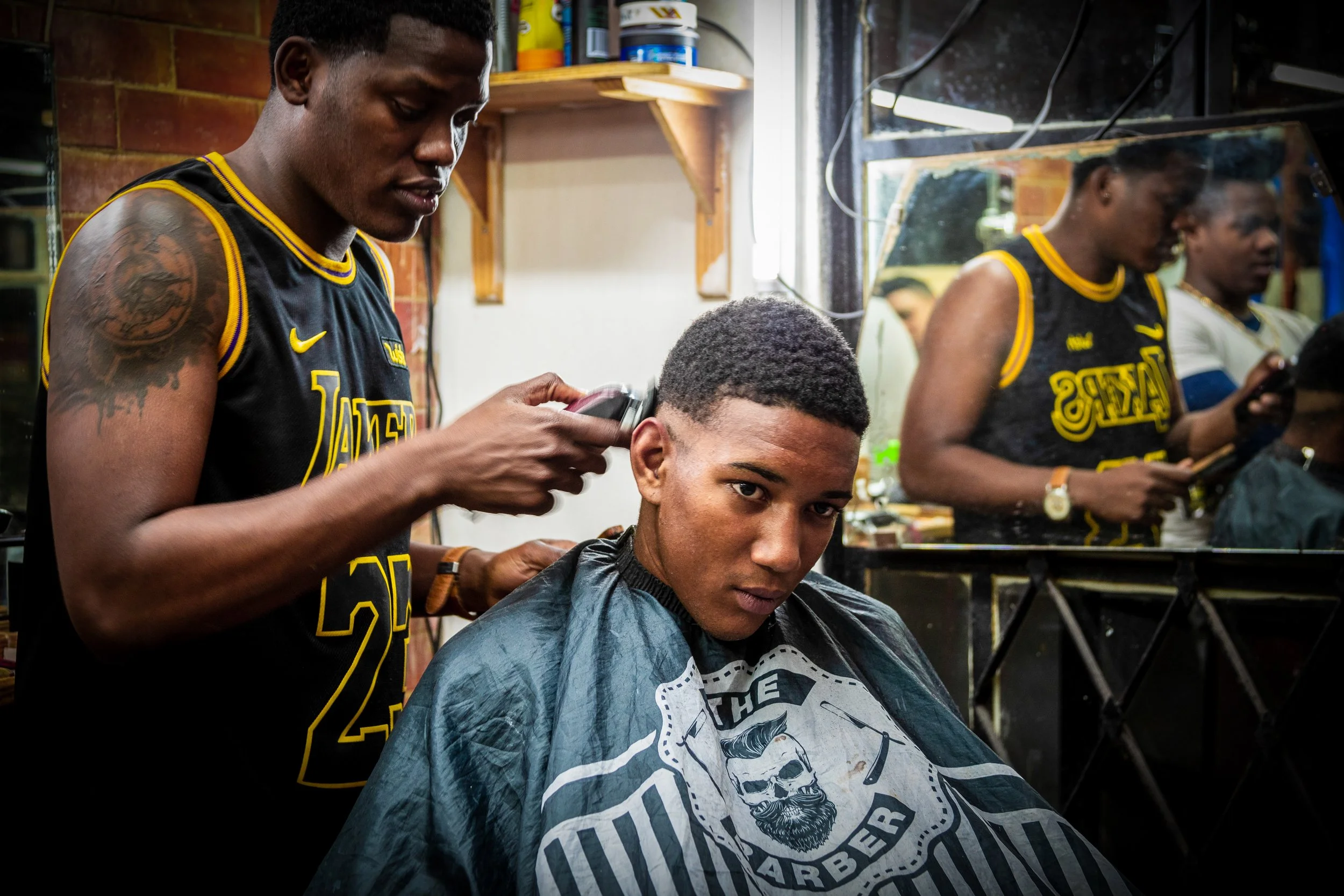 A young man getting a haircut at a barbershop with a barber using clippers. The client is wearing a barber cape with a skull logo. The barber is dressed in a black Lakers jersey. There is a large mirror behind the client reflecting the scene, and a s