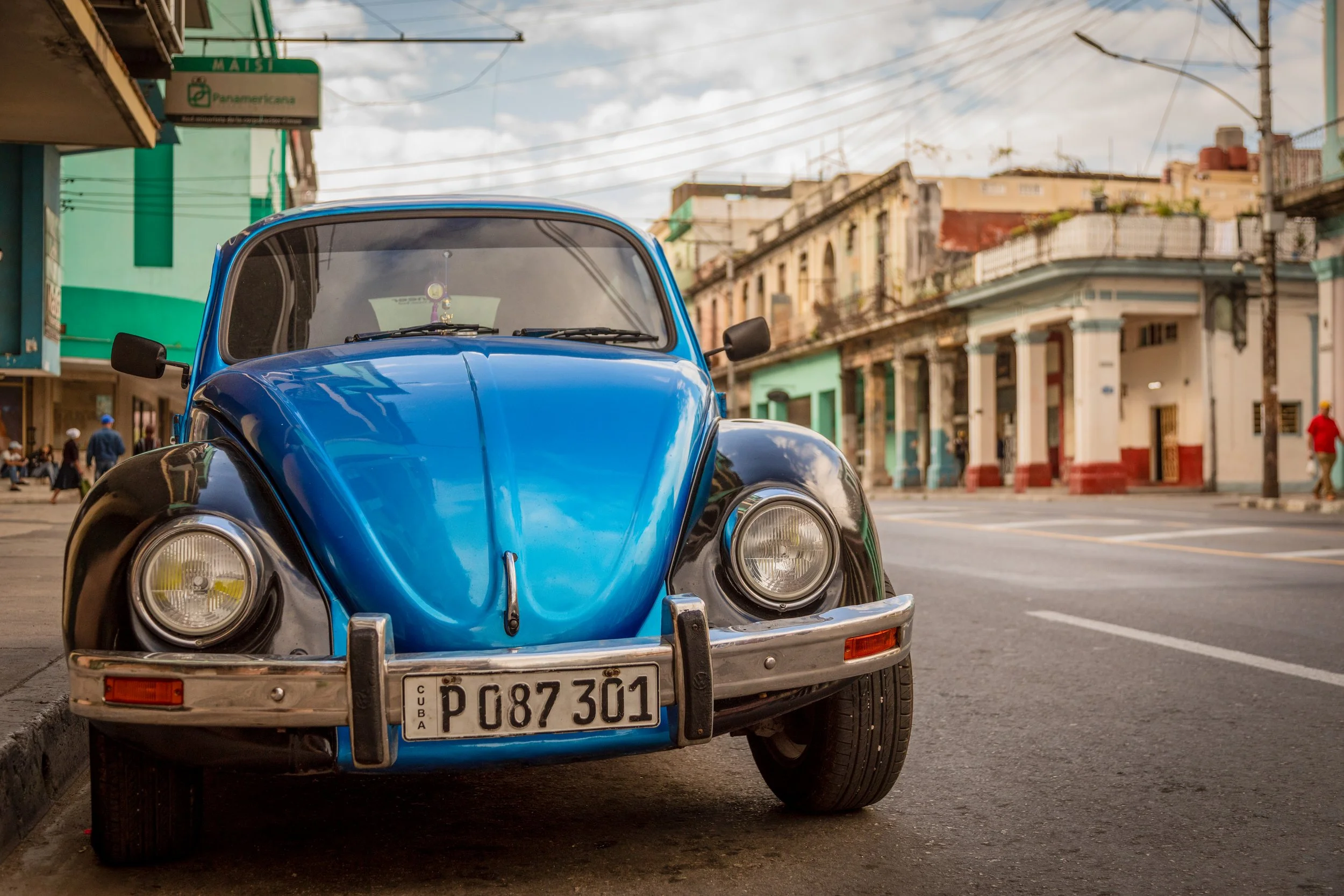 A vintage blue and black Volkswagen Beetle parked on a city street with colorful buildings and pedestrians in the background.