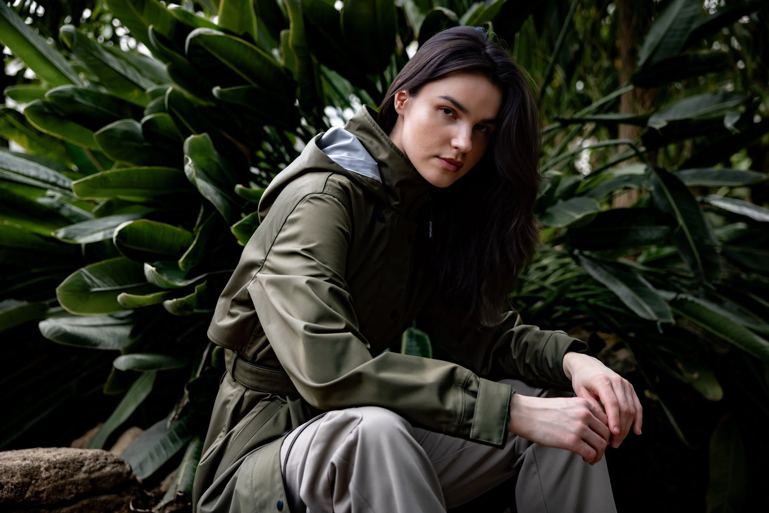 A woman with long dark hair in outdoor hiking gear, including a green jacket and light-colored pants, is sitting and looking at the camera amidst lush green foliage.