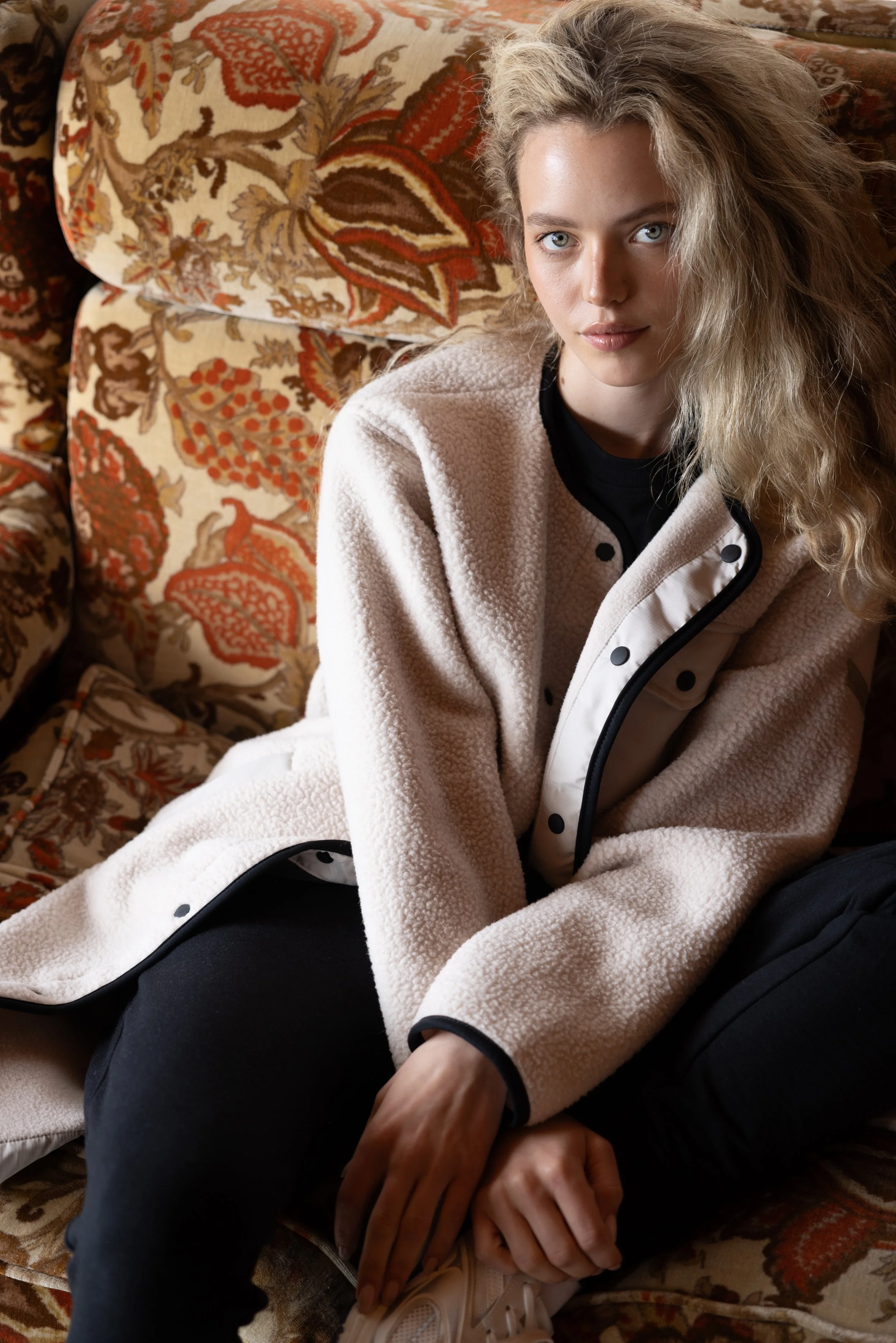 A young woman with wavy blonde hair and blue eyes sitting on a floral-patterned sofa, wearing a cream-colored fleece jacket over a black shirt and black pants.