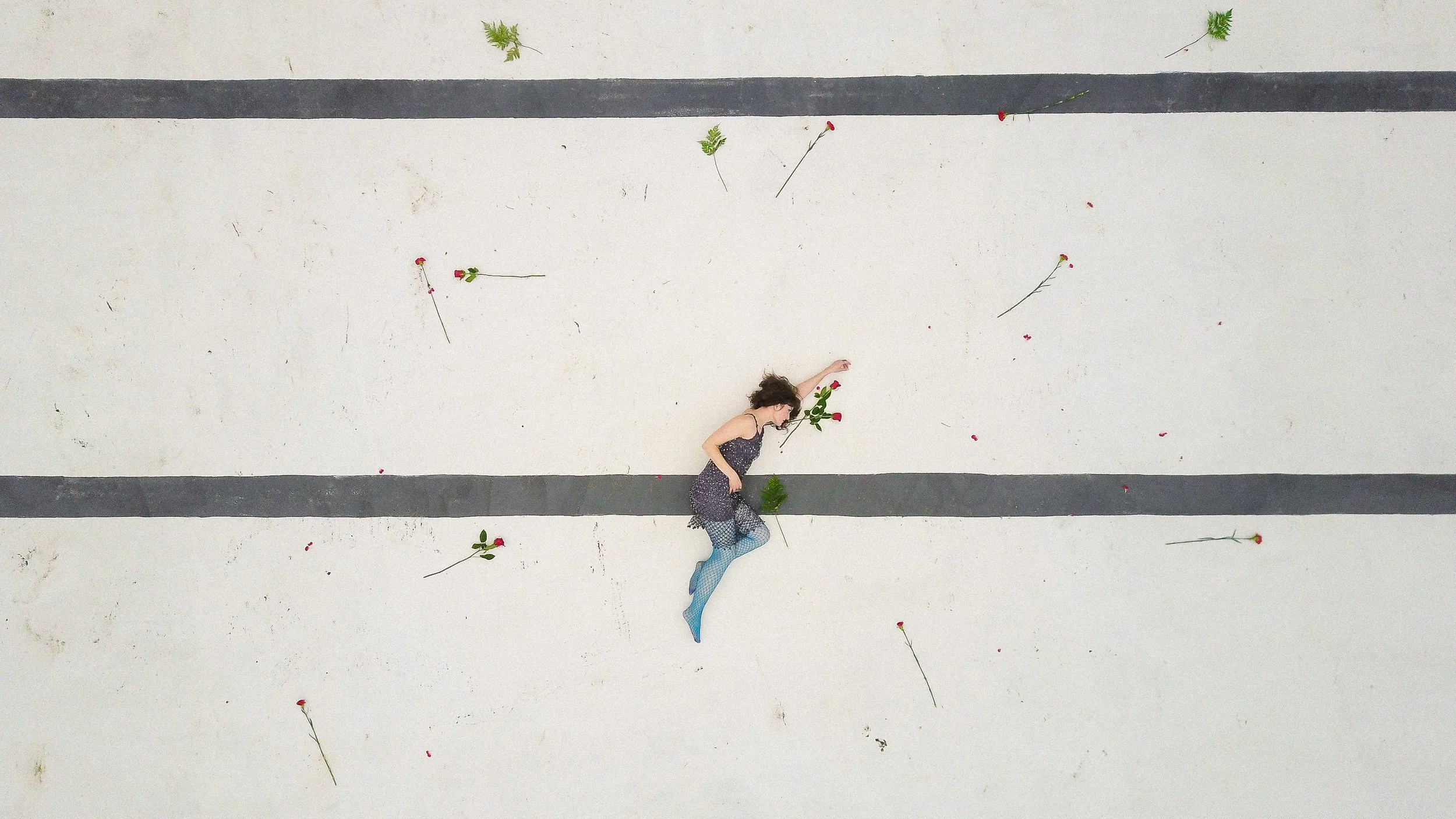 A woman lying on a white concrete surface with black lines, surrounded by scattered red roses and green leaves.