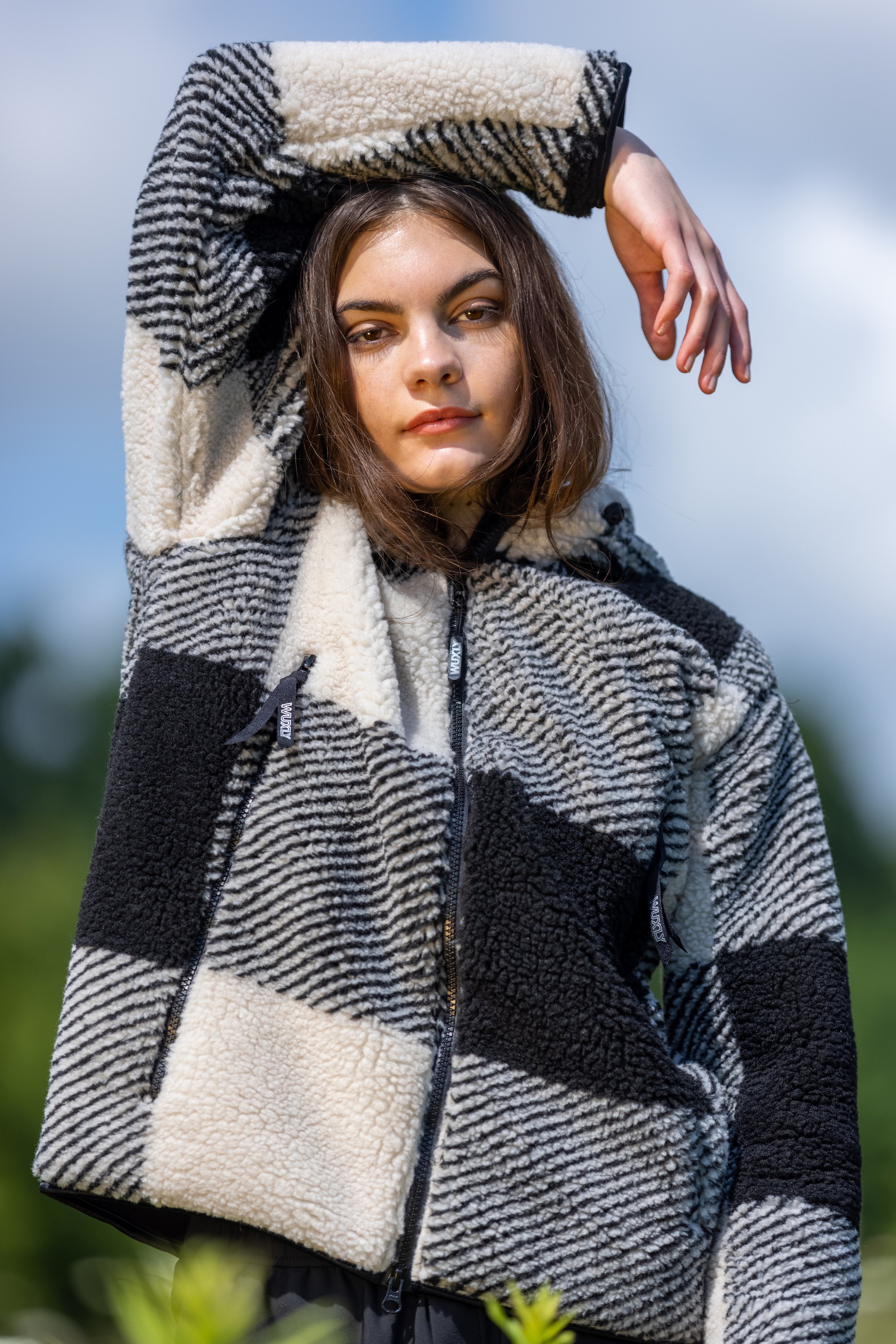 A young woman wearing a black and white patterned fleece jacket outdoors, with her right arm raised above her head and hand resting on her forehead.