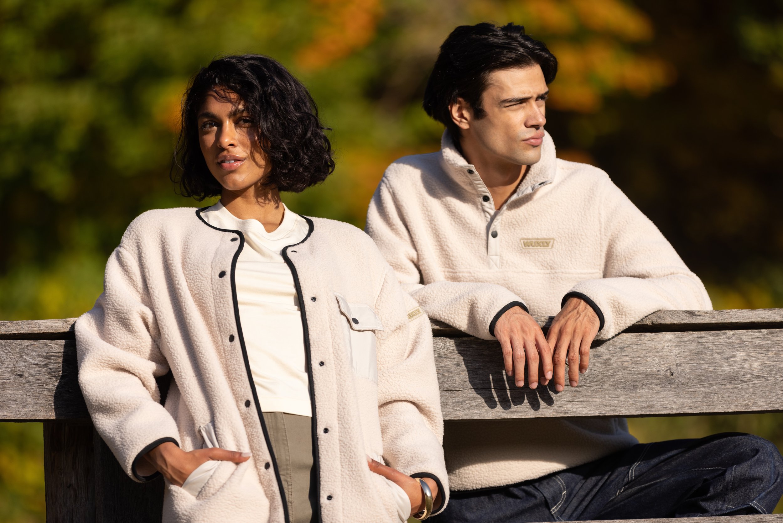 A woman and a man sitting on a wooden bench outdoors, wearing warm beige jackets, with a background of autumn foliage.