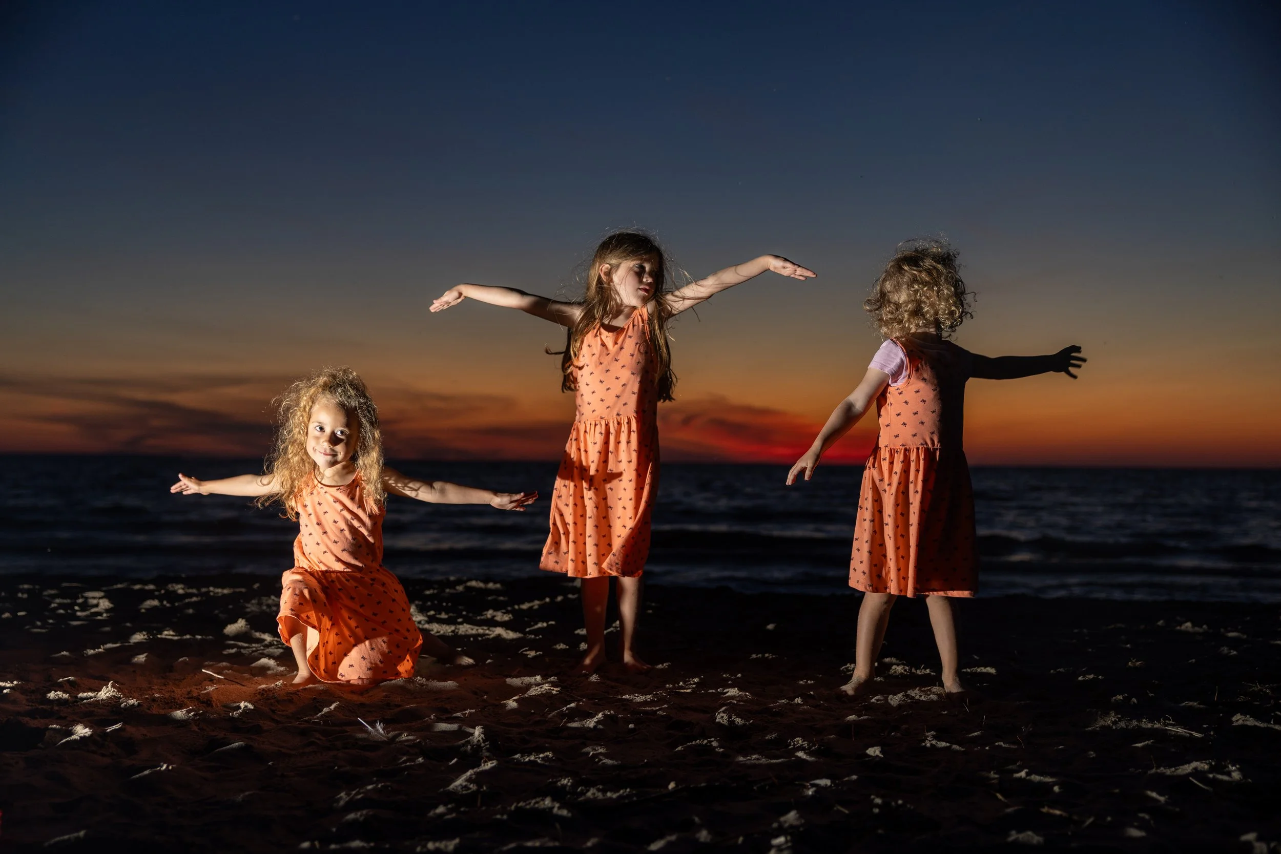 Three young girls in matching coral dresses with small black patterns playing on a beach during sunset. One girl is kneeling on the sand with outstretched arms, the second is standing with arms extended sideways, and the third girl stands with arms r