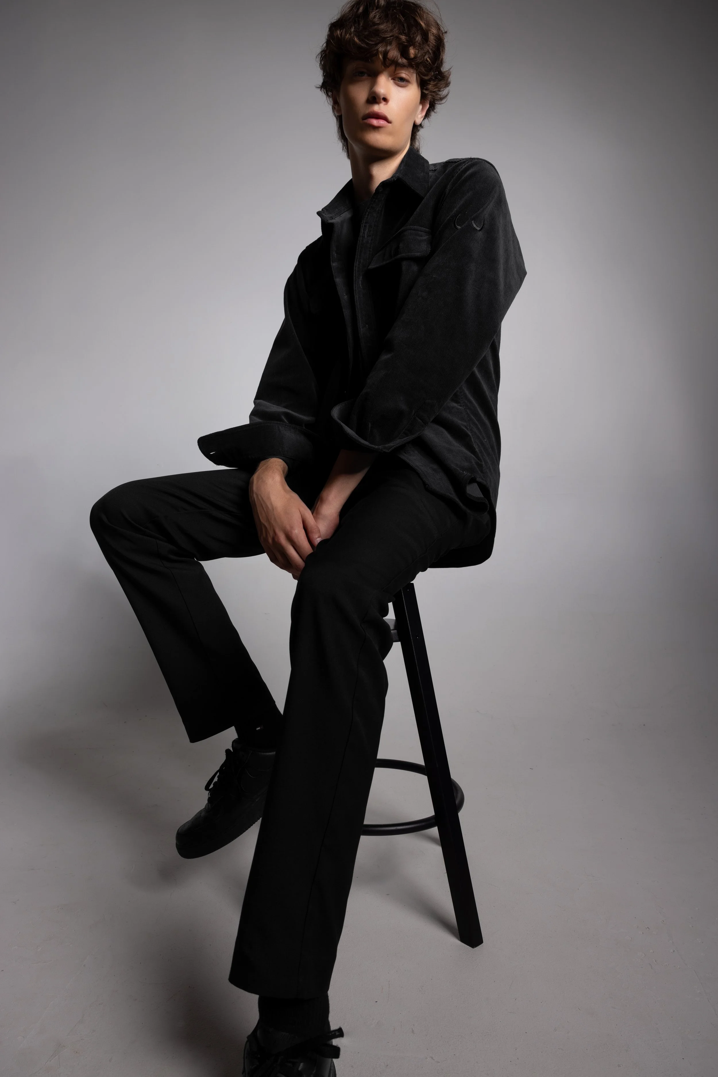 A young person with curly brown hair sitting on a stool against a plain gray background, wearing an all-black outfit.
