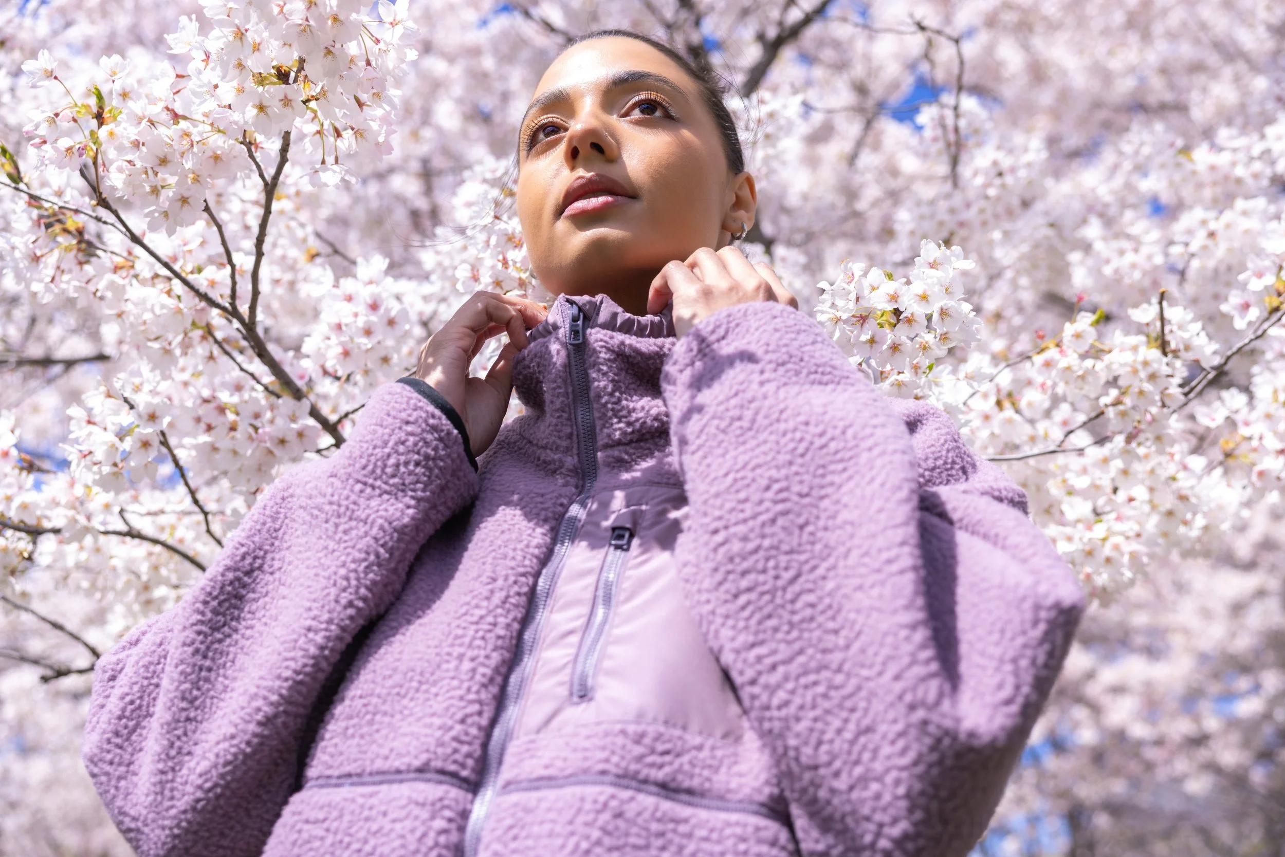 A woman adjusting her purple fleece jacket under blooming cherry blossom trees with pink and white flowers.