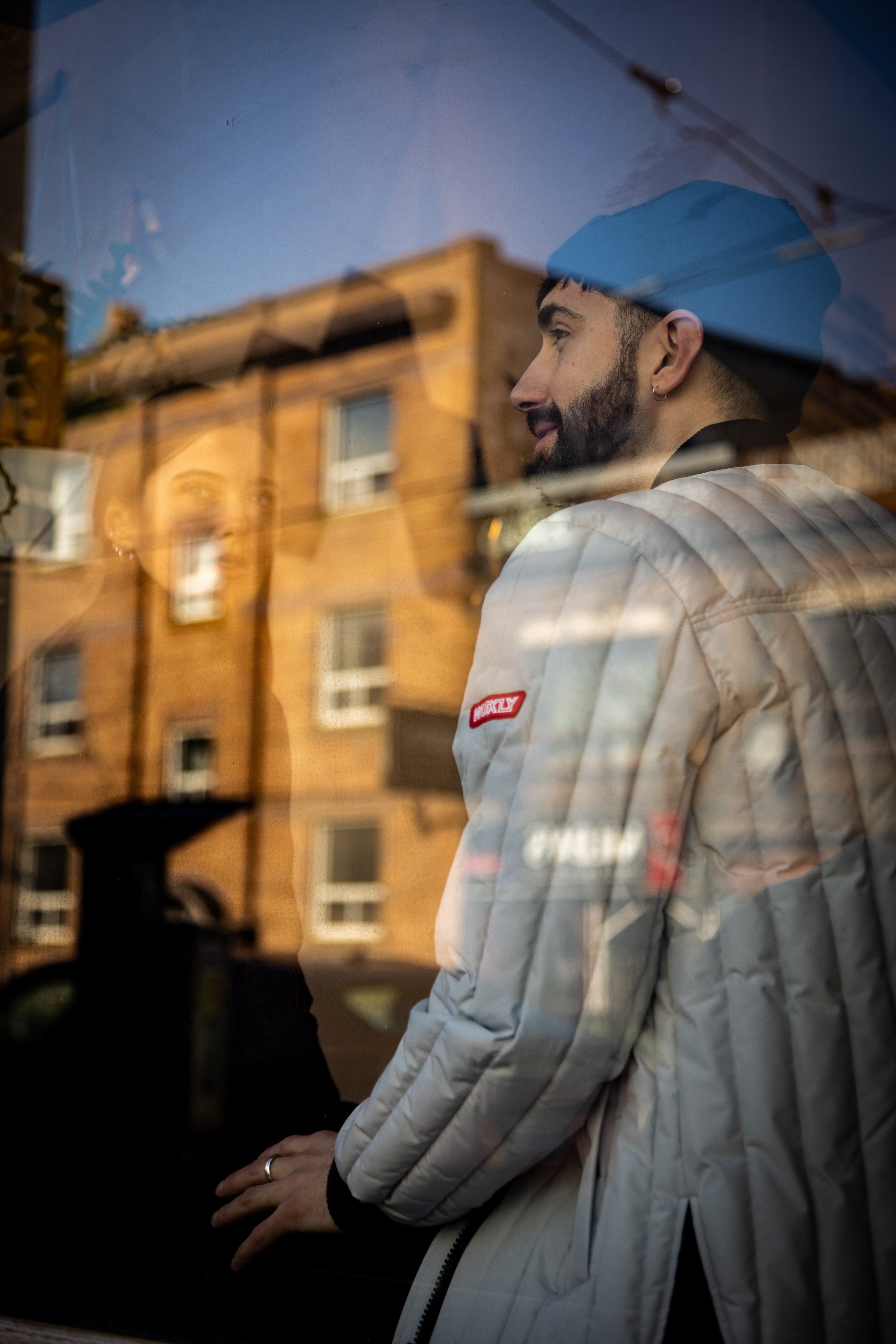 A man with a beard wearing a light-colored jacket and a dark cap, reflected in a window with a city building in the background.