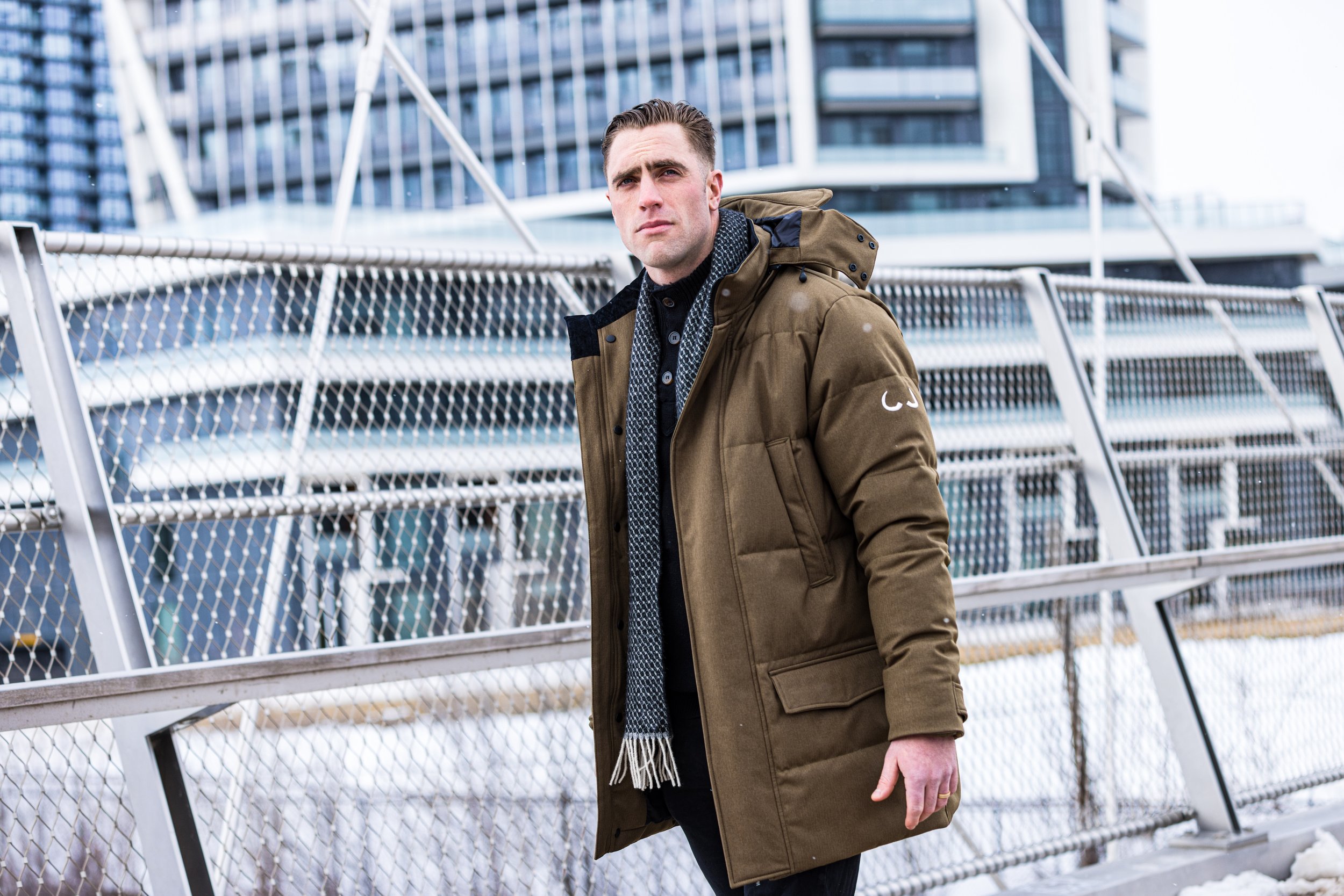 A man in a brown jacket with a hood standing outdoors near a metal fence in an urban setting with modern high-rise buildings.