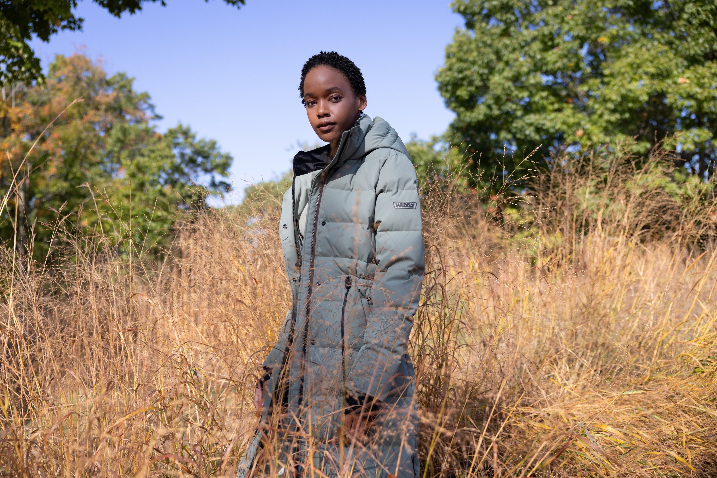 A woman standing in a field of tall, dry grass, wearing a light gray puffer jacket with a black hood, under clear blue skies and with green trees in the background.