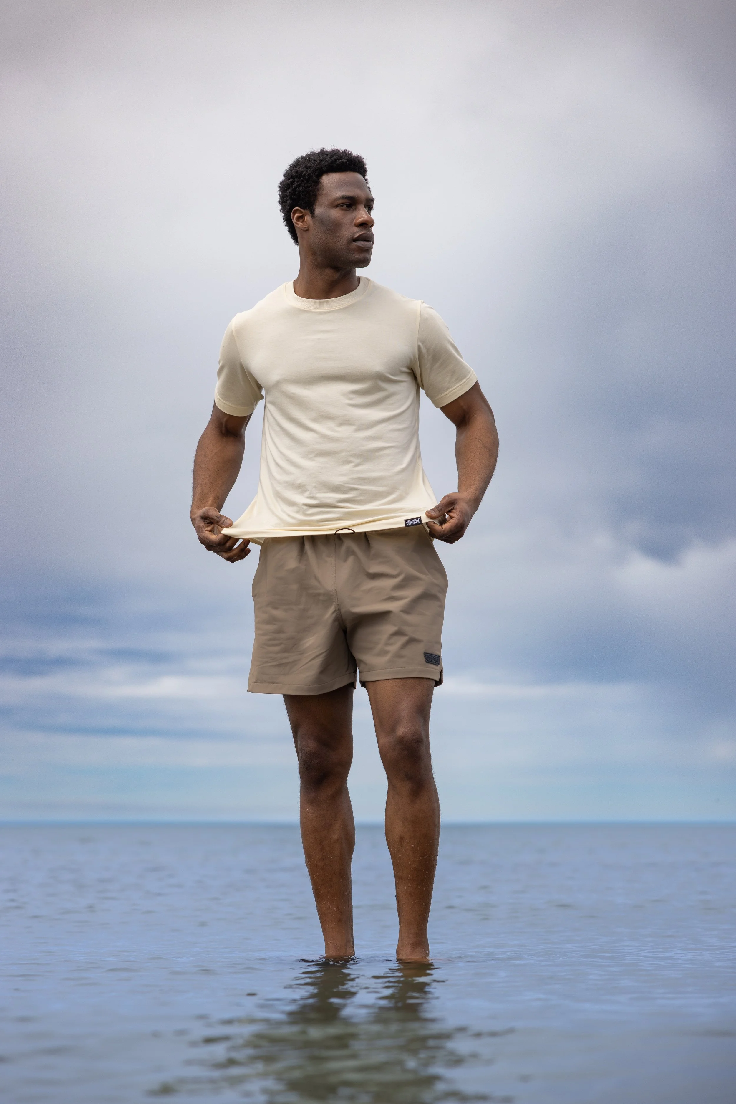 A man standing in shallow water at the beach, wearing a beige t-shirt and khaki shorts, with cloudy sky in the background.