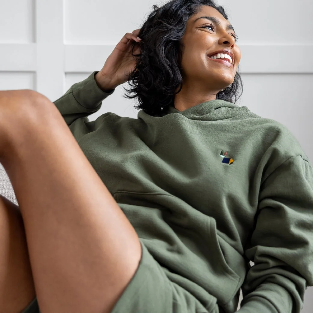 A woman with dark hair and a big smile, wearing a green hoodie and shorts, sitting comfortably and smiling against a white wall background.