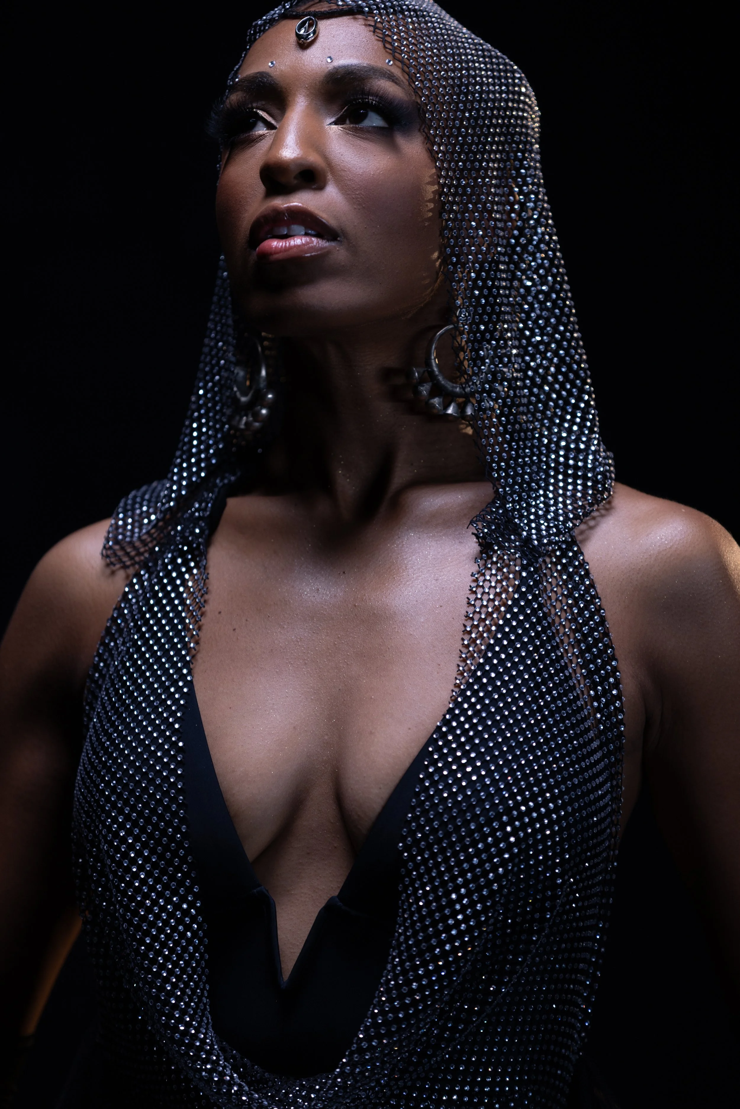 A woman wearing a rhinestone headpiece and earrings, with a dramatic gaze, dressed in a black, low-cut dress against a black background.
