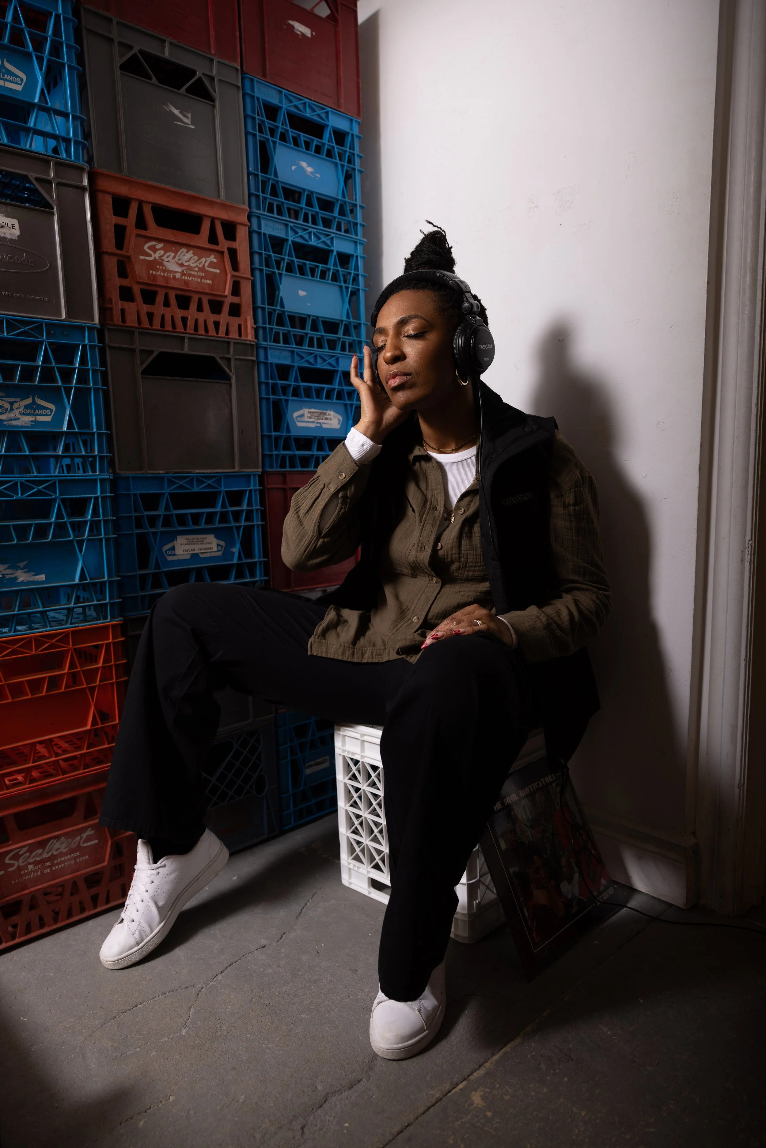 A woman sitting on a white plastic crate, wearing headphones, and listening to music in a room with stacked plastic crates against the wall.
