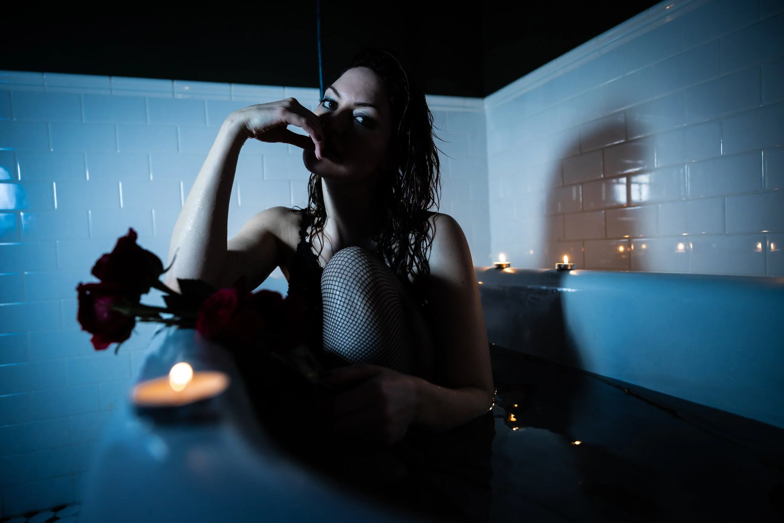 A woman with wet hair sits in a bathtub filled with water, surrounded by lit candles and a red rose, in a dimly lit room with white tiled walls.