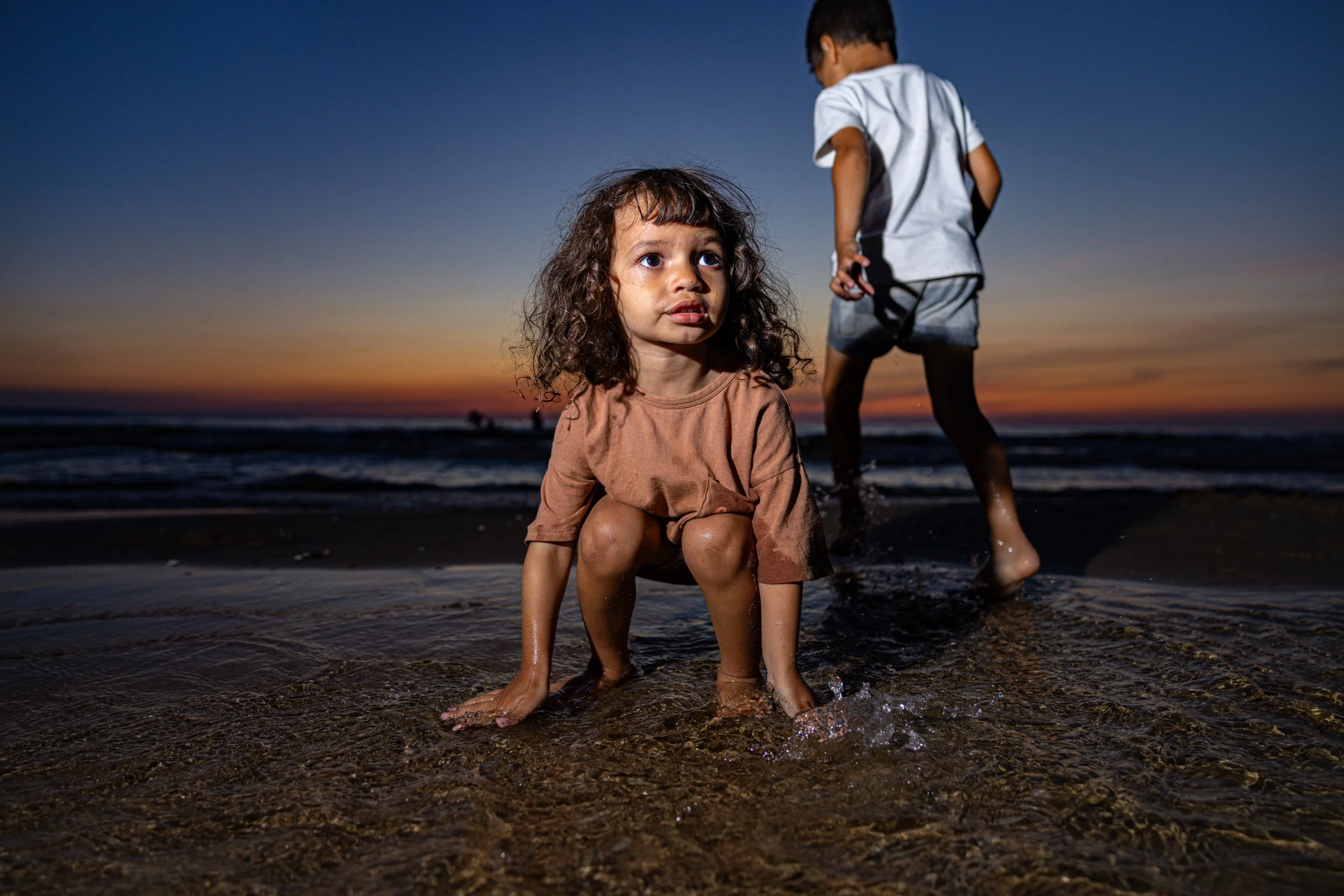 A young girl squatting on the beach during sunset, with a boy walking behind her as they play in the shallow water.