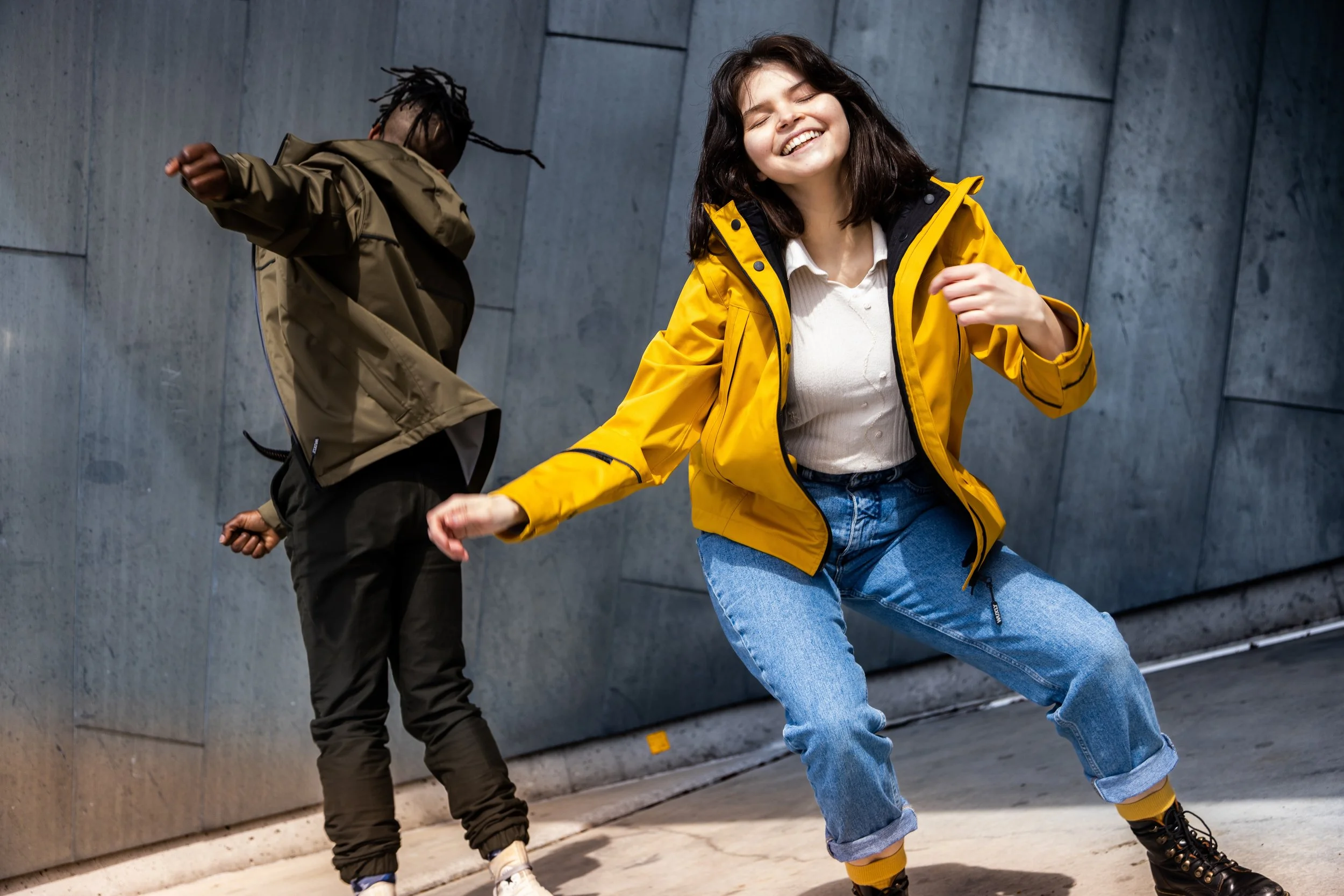 Two young adults smiling and dancing in front of a modern gray concrete wall, dressed casually in jackets and jeans.