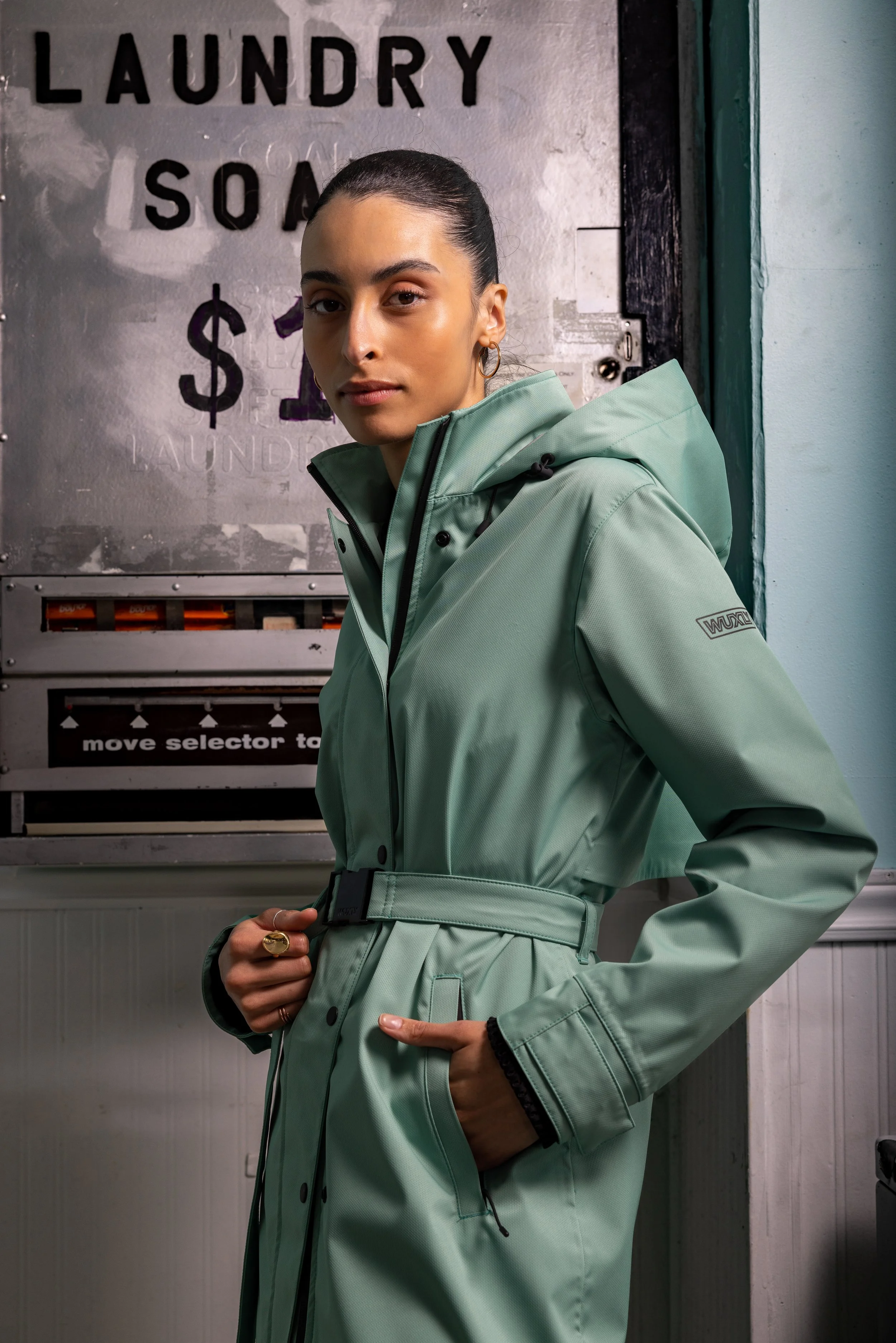 A woman in a light green raincoat standing in front of a laundry soap vending machine.