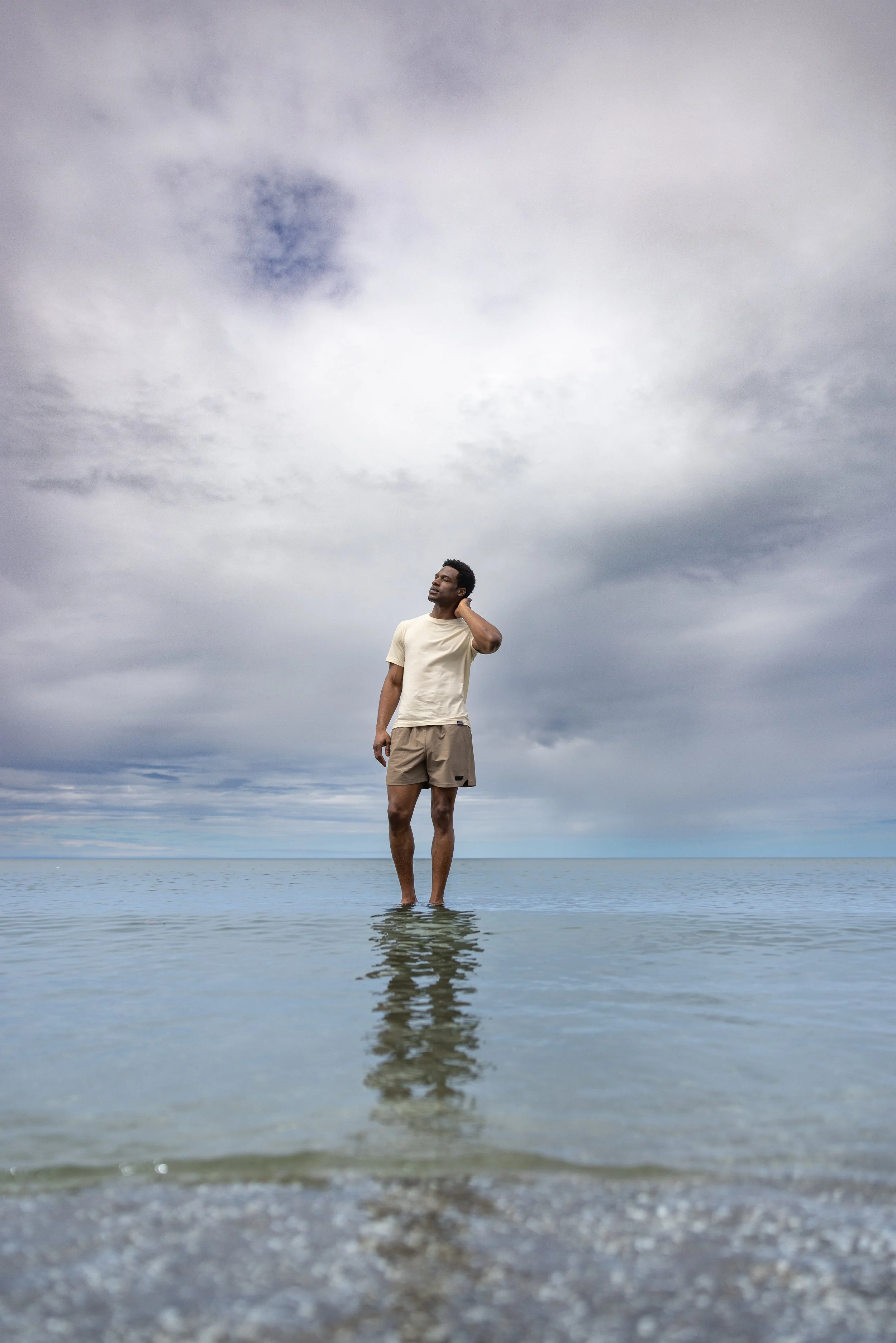 A man standing in shallow water at the beach with a cloudy sky behind him.