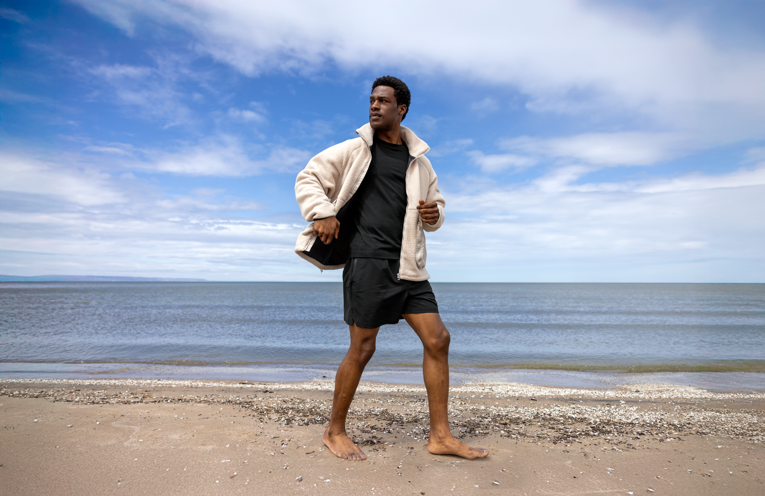 A man jogging barefoot on a beach with sand and shells, wearing a black T-shirt, black shorts, and a beige fleece jacket, with an ocean and partly cloudy sky in the background.