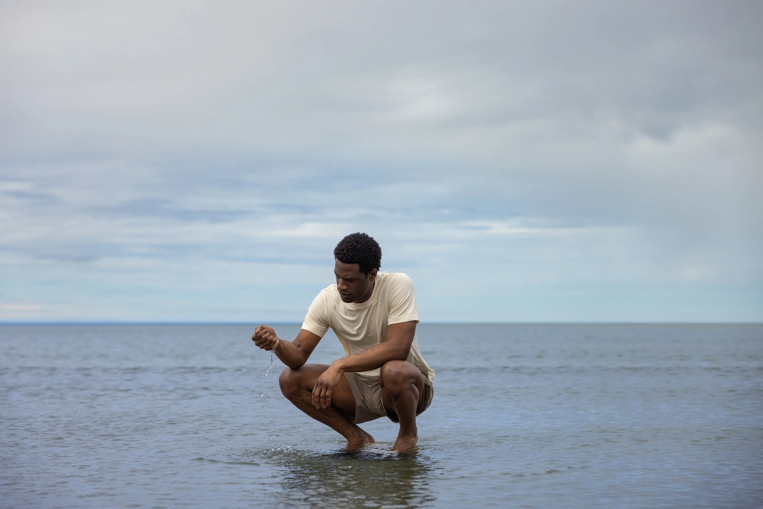 A man crouching in shallow water at the beach, splashing water with his hand on a cloudy day.