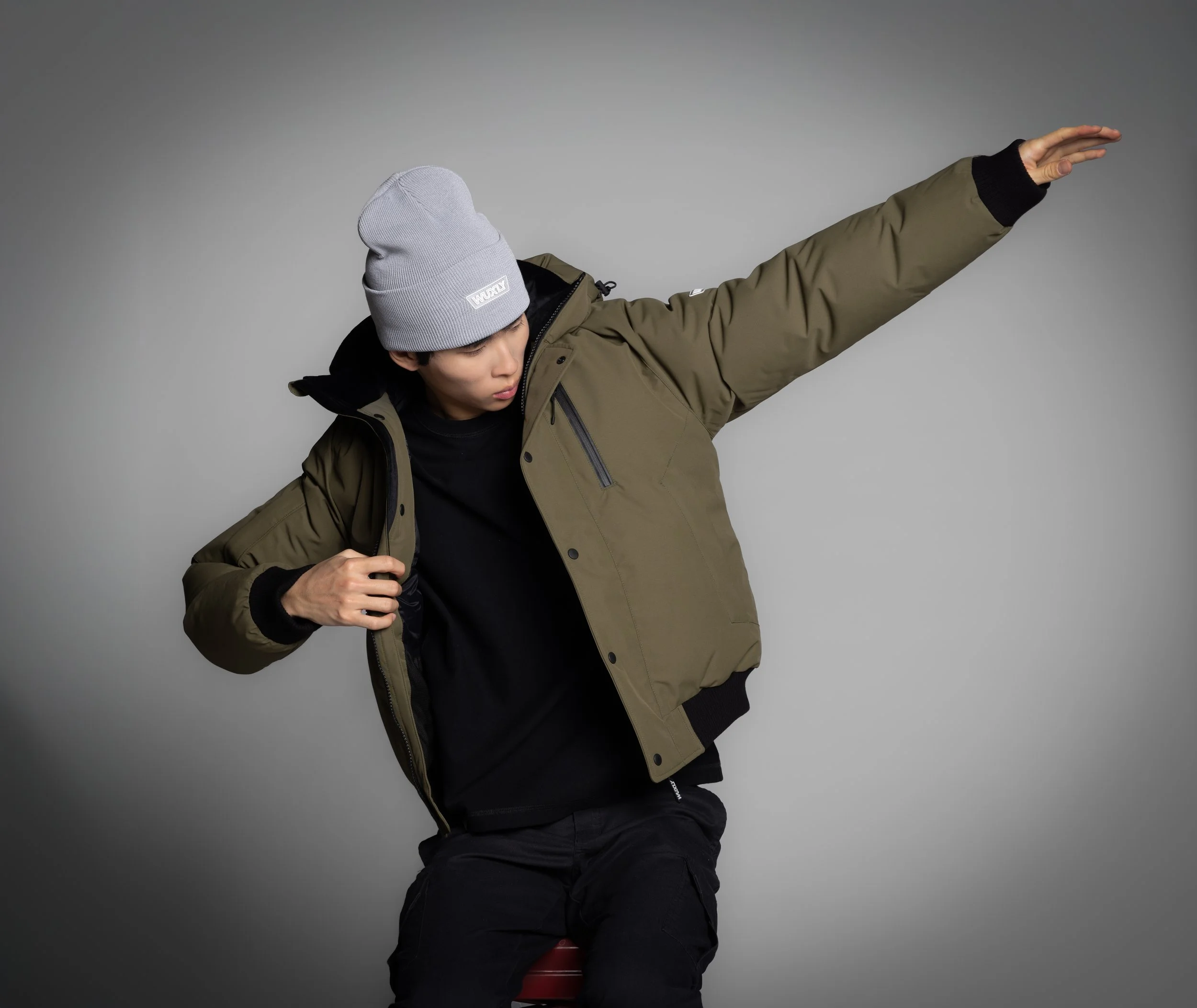 A young man wearing a gray beanie, black t-shirt, and an olive green jacket, sitting on a red chair. He is adjusting his jacket with one hand and extending his other arm to the side, against a gray background.