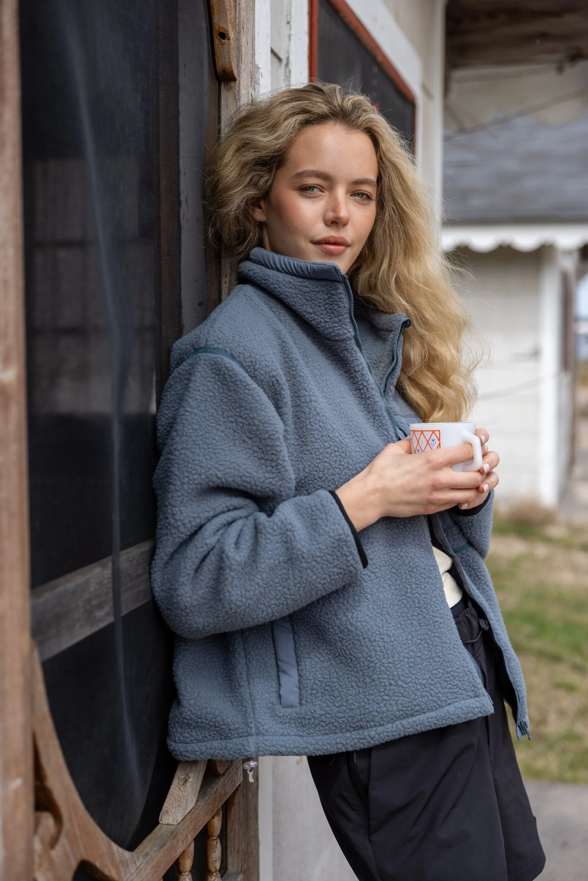 A young woman with long, wavy blonde hair wearing a blue fleece jacket and black pants, holding a cup, standing outdoors against a rustic wooden wall.